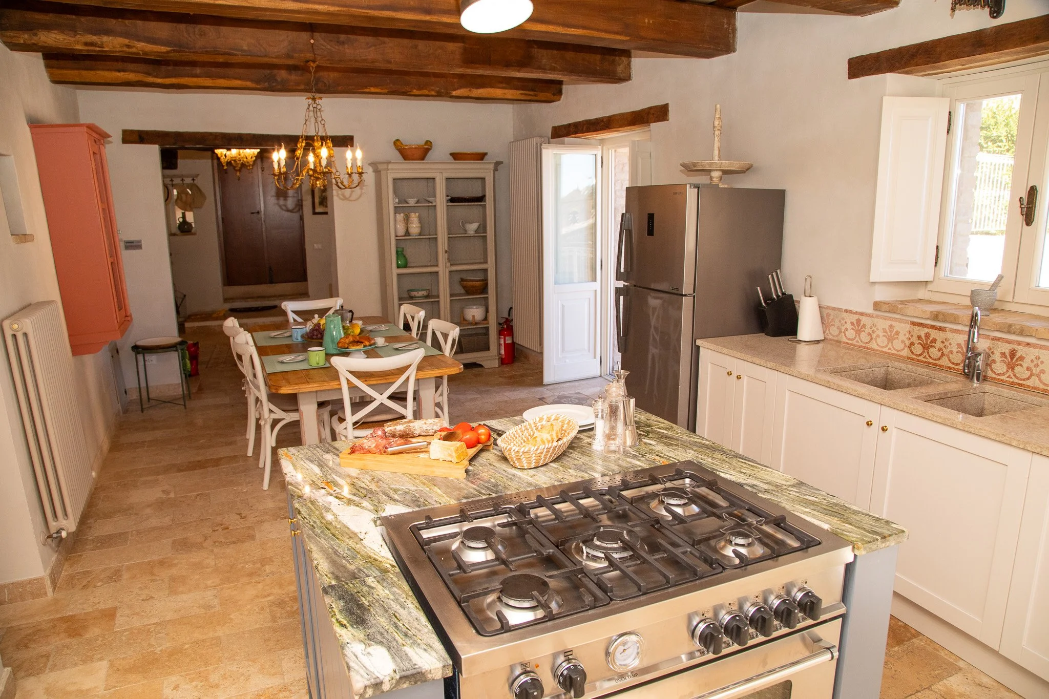 Rustic kitchen with wooden ceiling beams, a marble countertop gas stove, white cabinets, and a dining table with chairs in the background, decorated with dishes and food. Il Riposo, Patrignone, Le Marche, Appassionata, Fractional Ownership