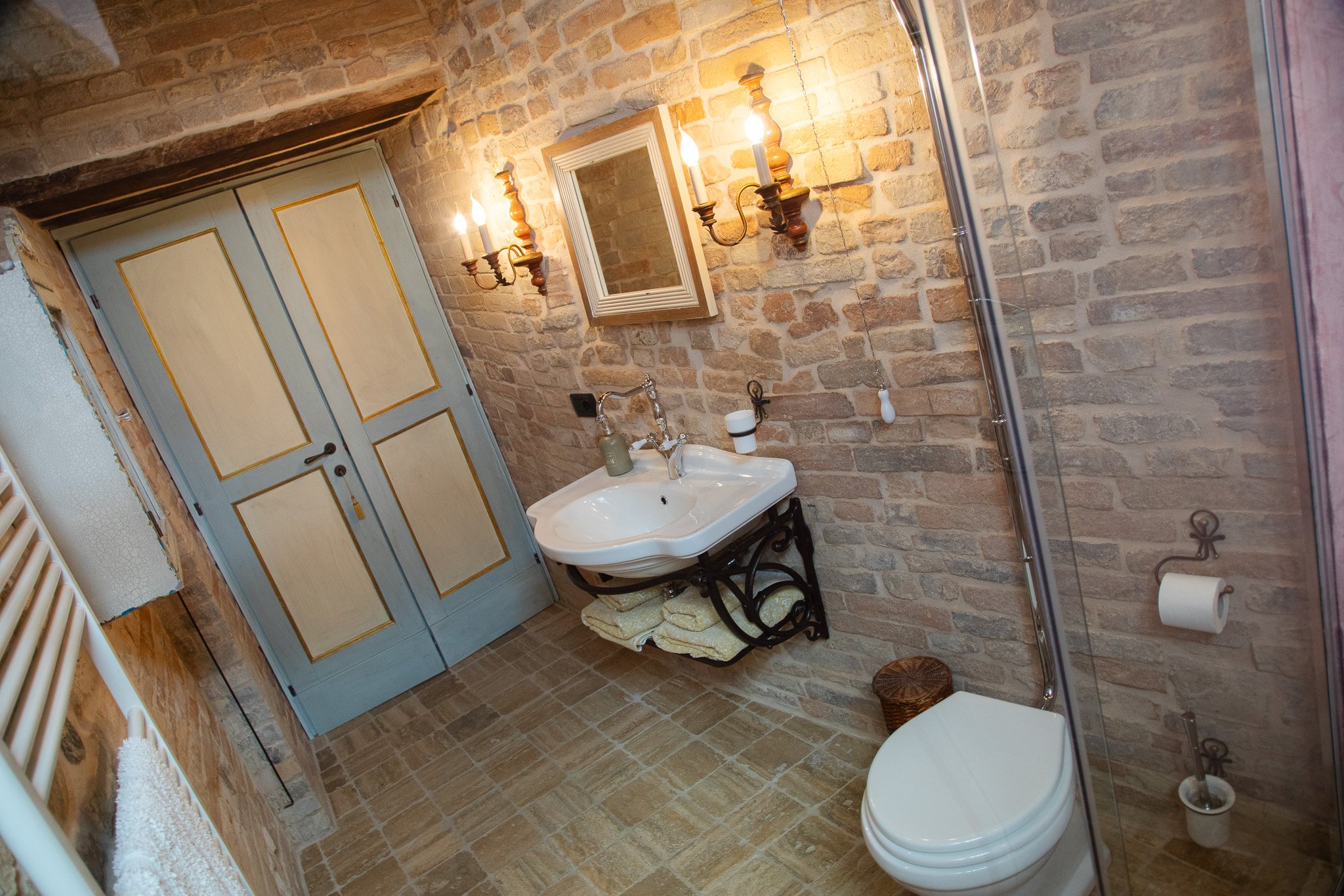 Rustic bathroom with exposed brick walls, a white sink, mirror, vintage-style light fixtures, wooden door, and white toilet. Il Nido Apartment, Petritoli, Appassionata Fractional Ownership.