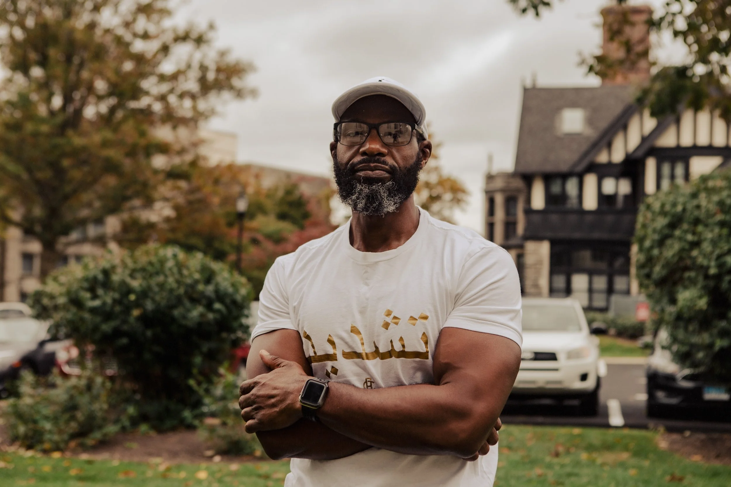 This image is of Stacey Torrance. He is standing with his arms crossed and is backed by grass. a parking lot, and a building. He is wearing a white shirt with gold Arabic writing, an apple watch, glasses, and a white hat.
