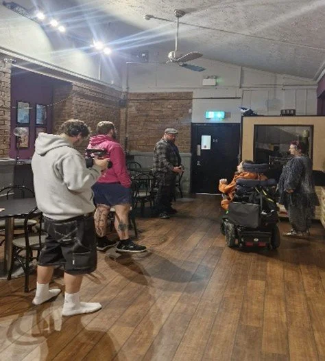 Four people talking in a circle with a someone filming there conversation. this space has a wooden floor, white celling and brick walls. There chairs and posters on the wall