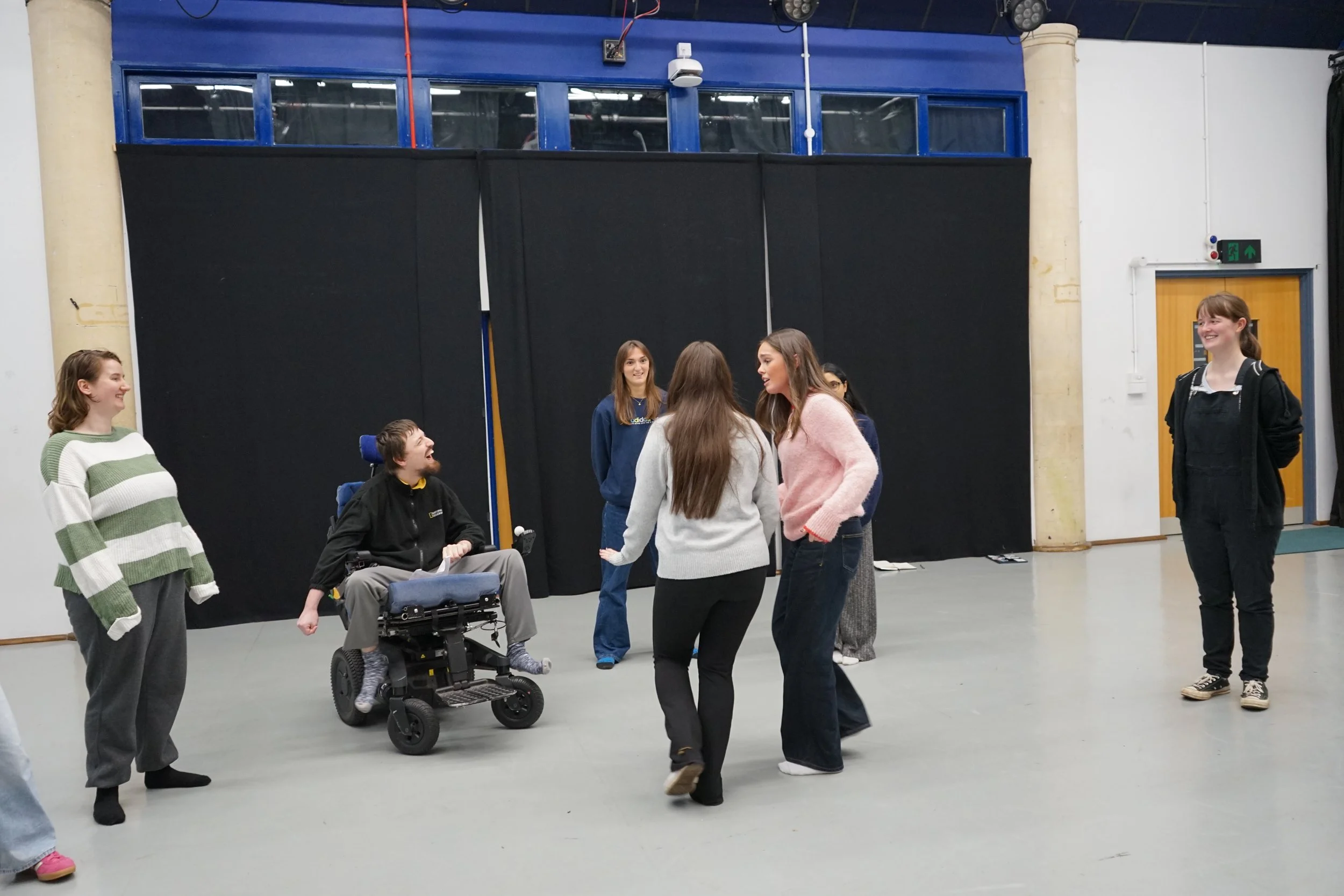 Three young students standing in a cluster looking at each other articulating. Others around them watch and laugh at the scene playing out.