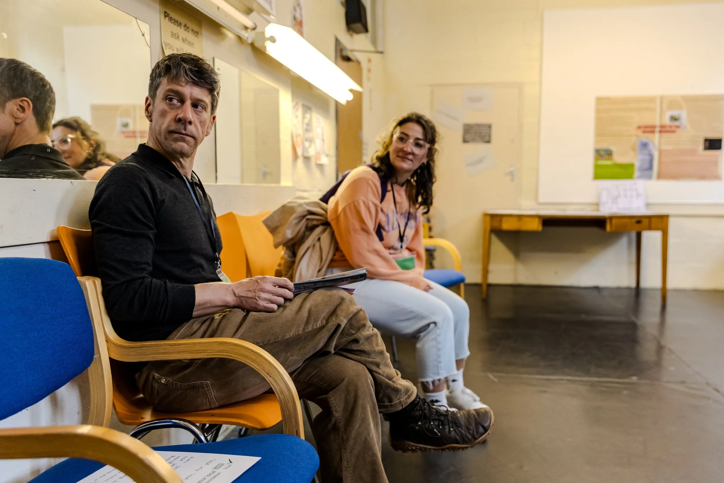 Two audience member peer around the corner from their chairs. Cross between mild intrigue and slight apprehension. The walls are yellow with posters everywhere.