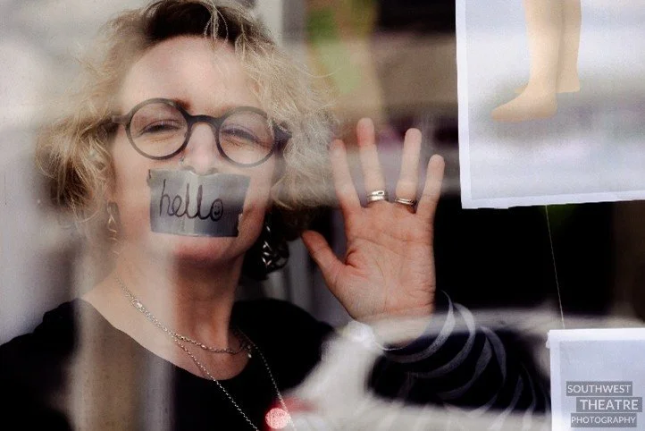 A woman in glasses with curly blonde hair waves at the camera. She has duct tape across her mouth with the word ‘hello’ written on it. Her eyes look smiley and there is a smiley face in the ‘O’.