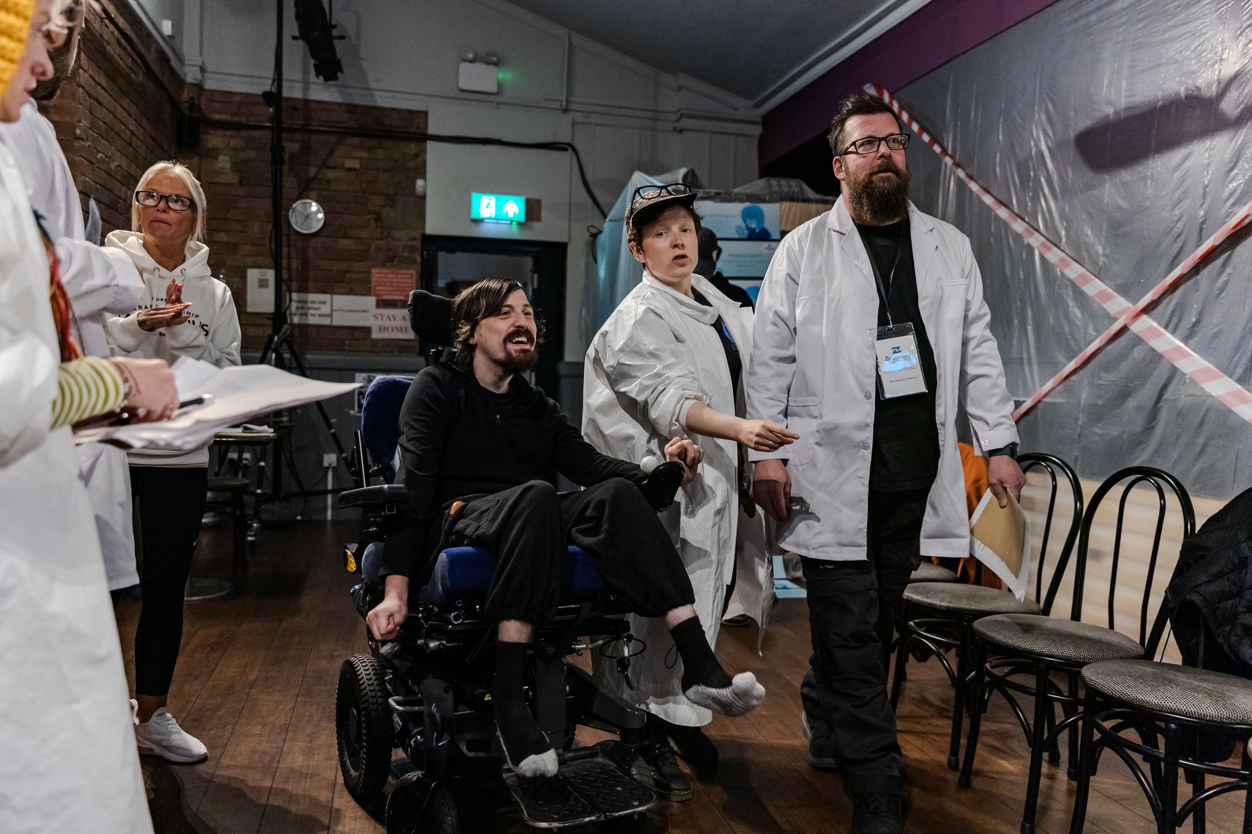 four performers in white lab coats and one in a casual jumper look pre ocoupied whilst the wheelchair users in black with messy brown hair has a wry smile