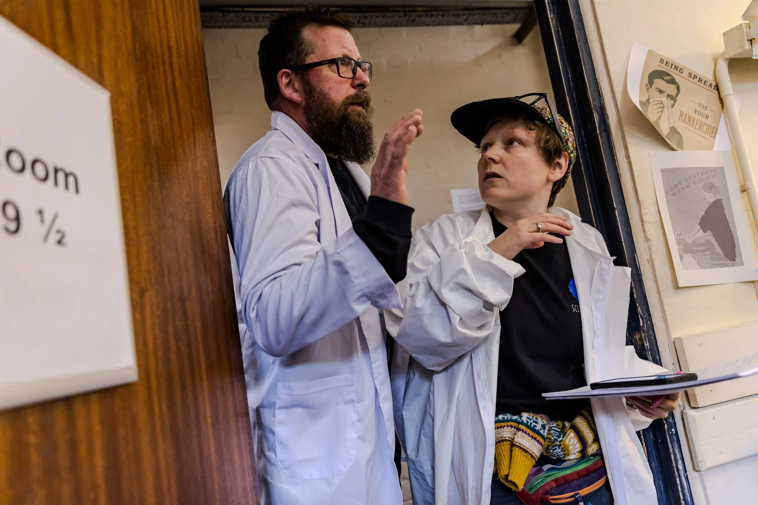 Two characters in white coats, barge through the door. One looks feverish around the room the other stare back with worry.