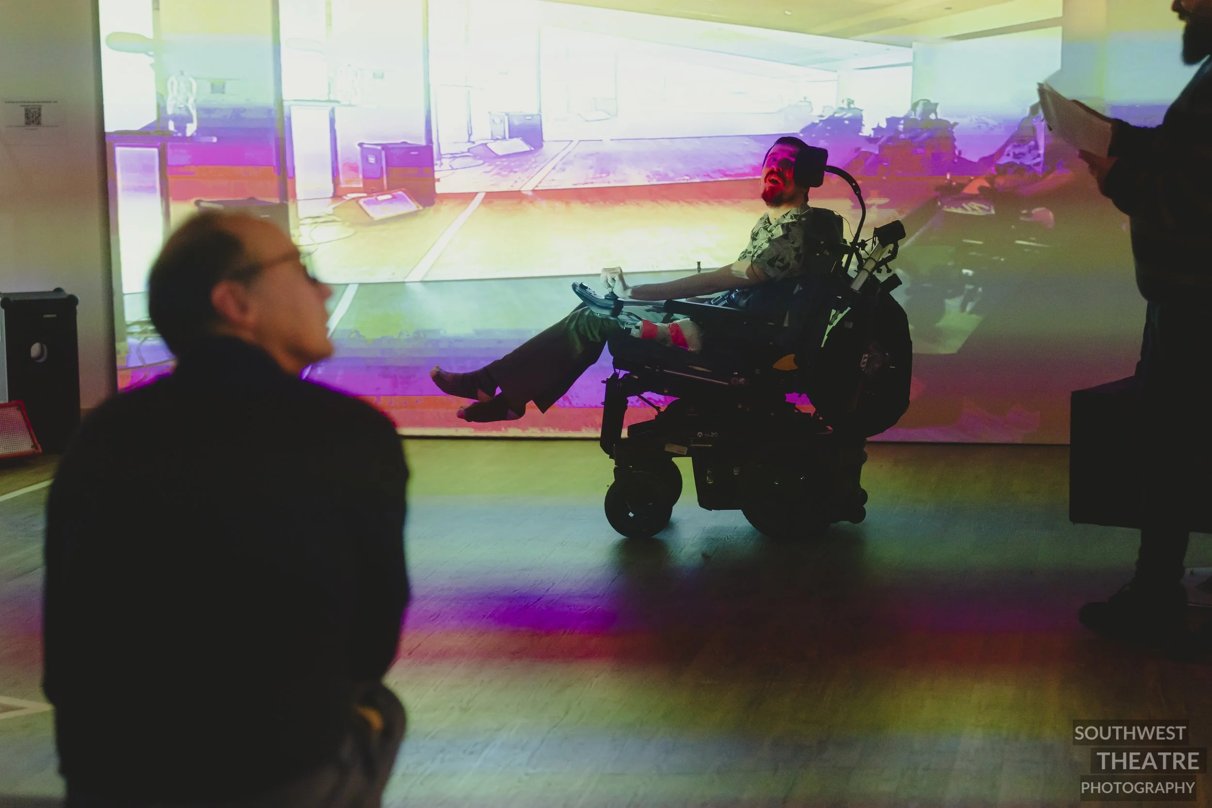 Audience member with glasses and brown hair looks questionably at a performer with a clip board. Middle, wheelchair-user shouts over their shoulder. The scene is replicated on a projection wall.