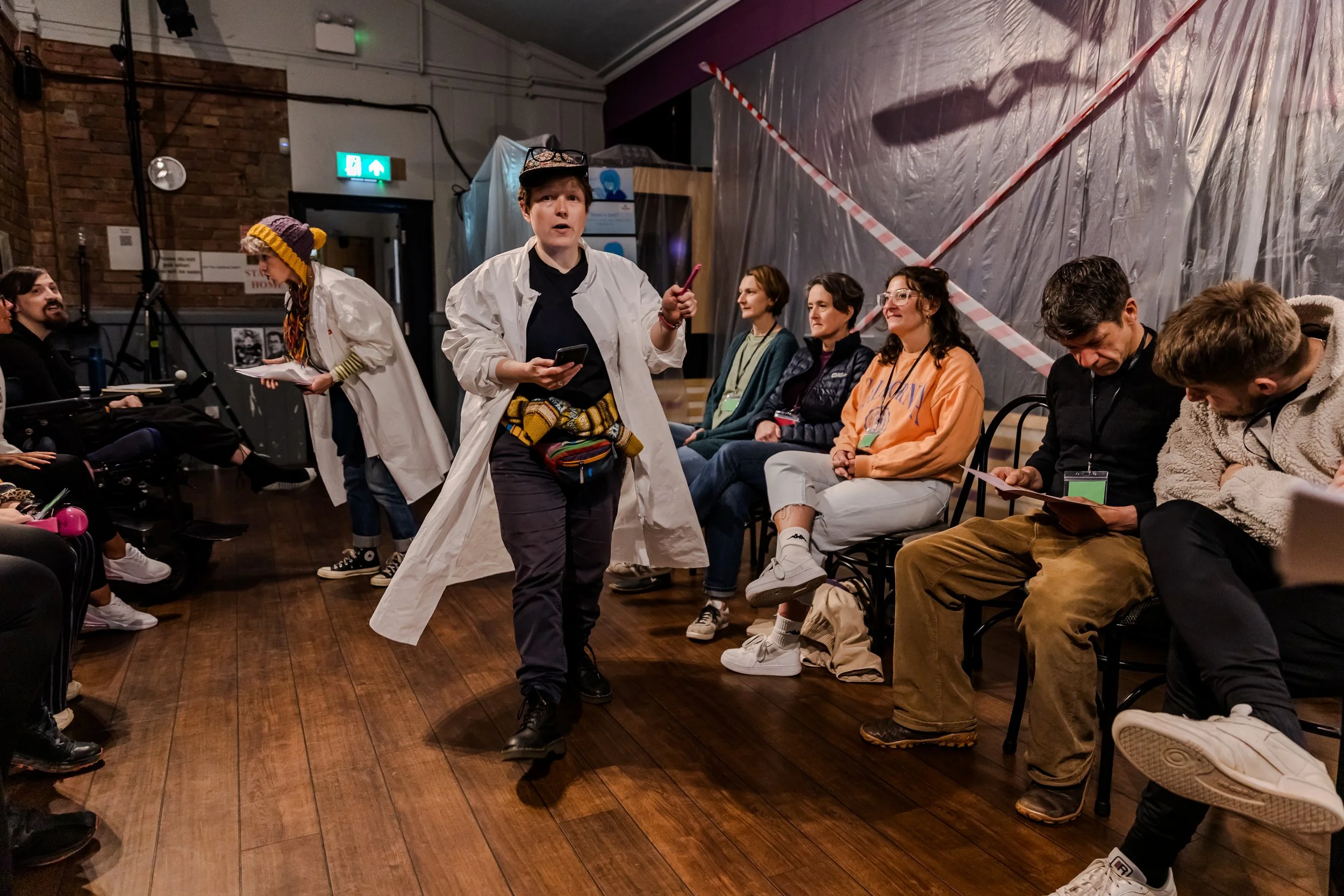 Busy waiting room out with someone in white lab coat rushing through. behind, second white coat performer leans over a wheelchair user with a clip board