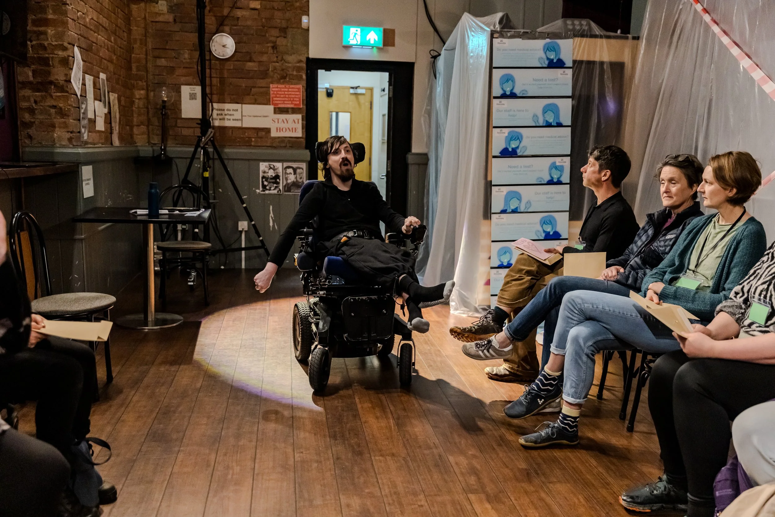 Busy wating room with a performer in black with messy hair delivers a speech in a shallow spot light