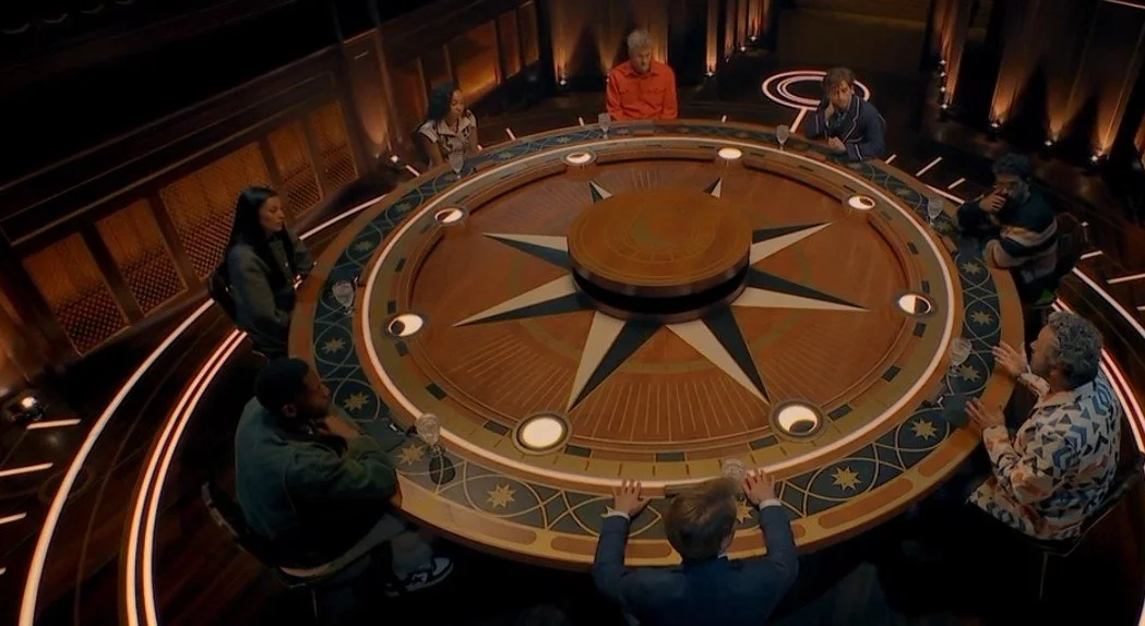 8 celebrities sitting around a grand wooden table, with moon and sun patterns decorating it.