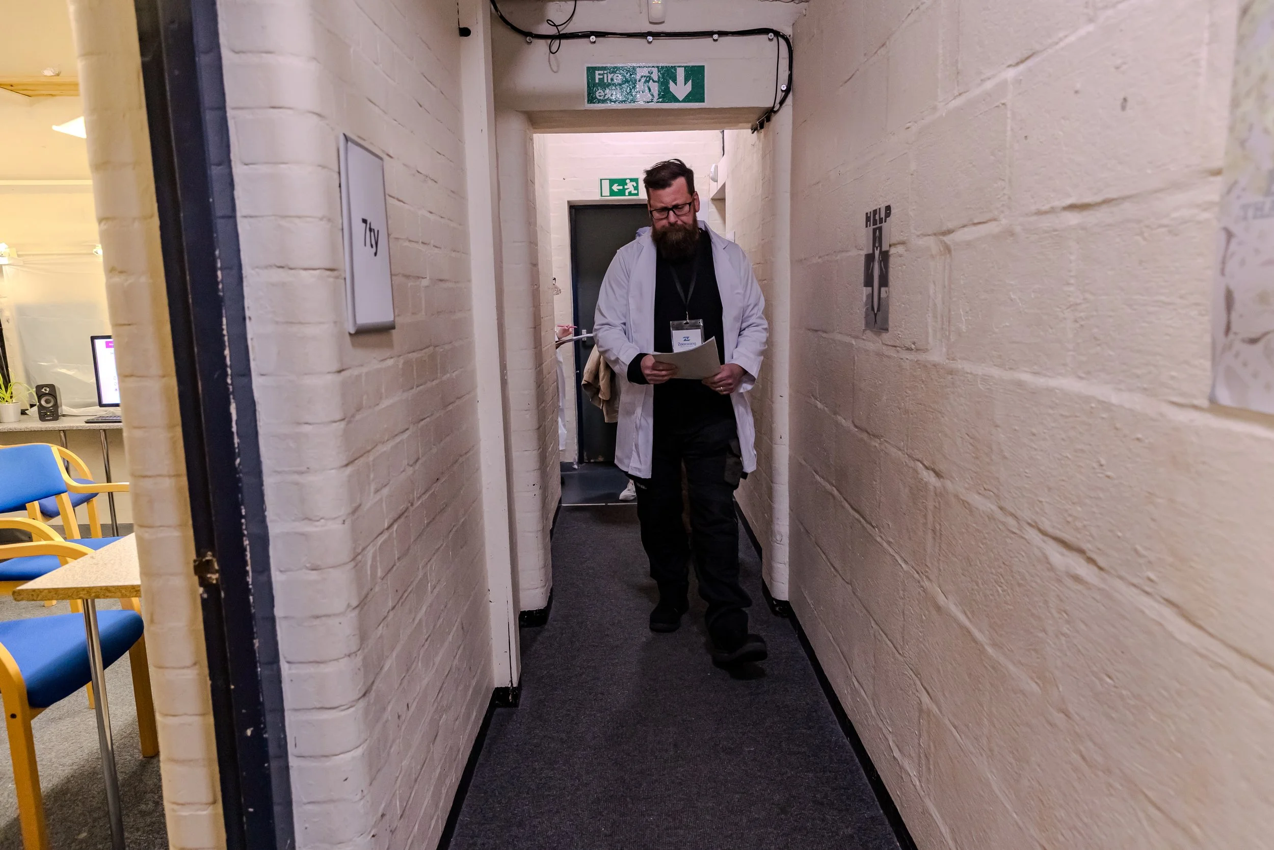 short corridor with white walls and a fire escape at the end. Left, a door left ajar showing a bright room with chairs and a screen. Walking towards the camera, the performer with a lab coat and long brown beard looks confused at paper in their hands