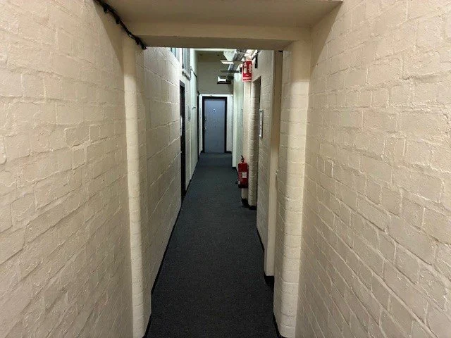 Long corridor with yellow brick walls and grey carpet. The door at the far end is shut.