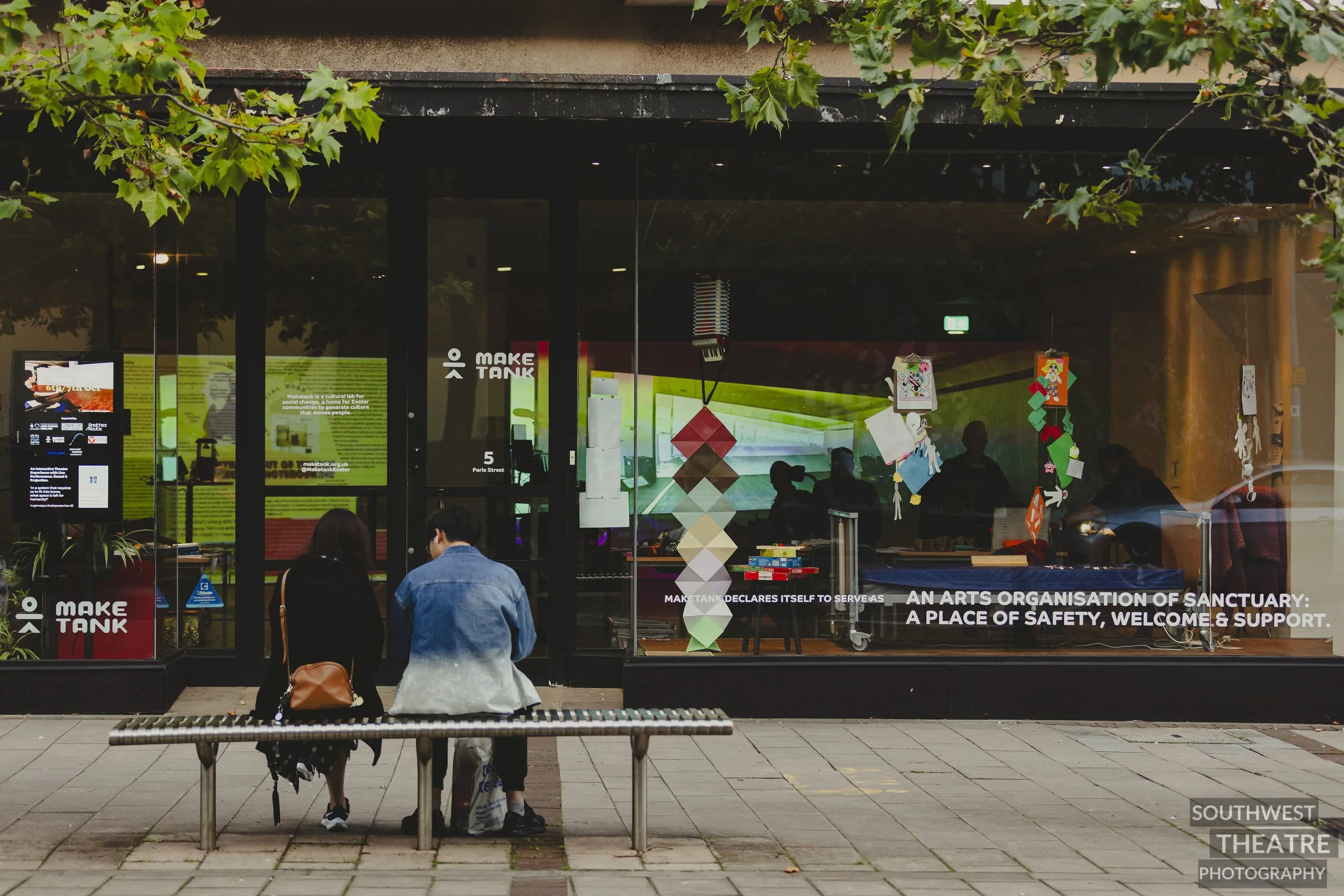 Two people on bench in front of a art shop window full of colour and set 