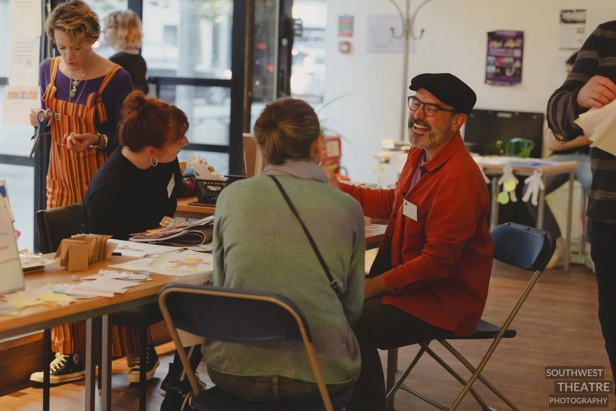 three people around a messy creative table interacting with each other. A tall women with a purple top and orange dungarees focusing on scissors and tape on her hands.