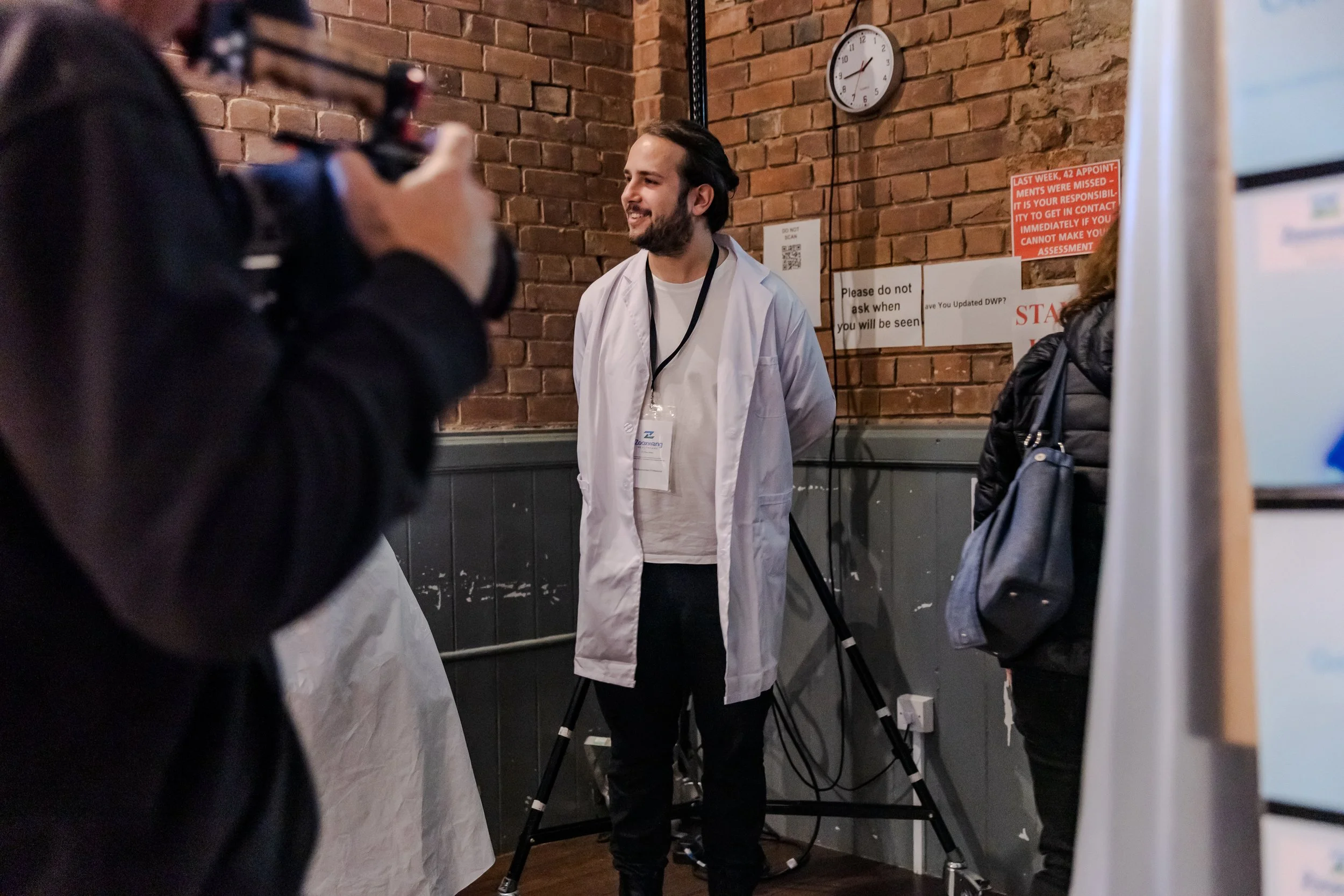 performer in while lab coat and brown hair stands Infront of brick wall with posters. Left camera operator