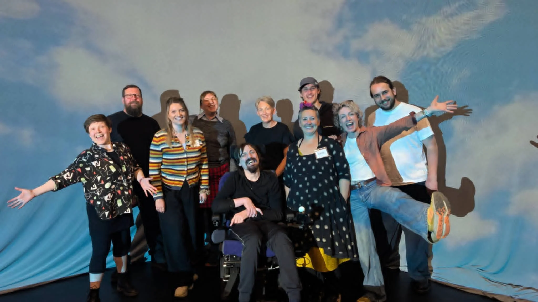 Ten people grin in a group photo with a large projection of fluffy clouds behind them.