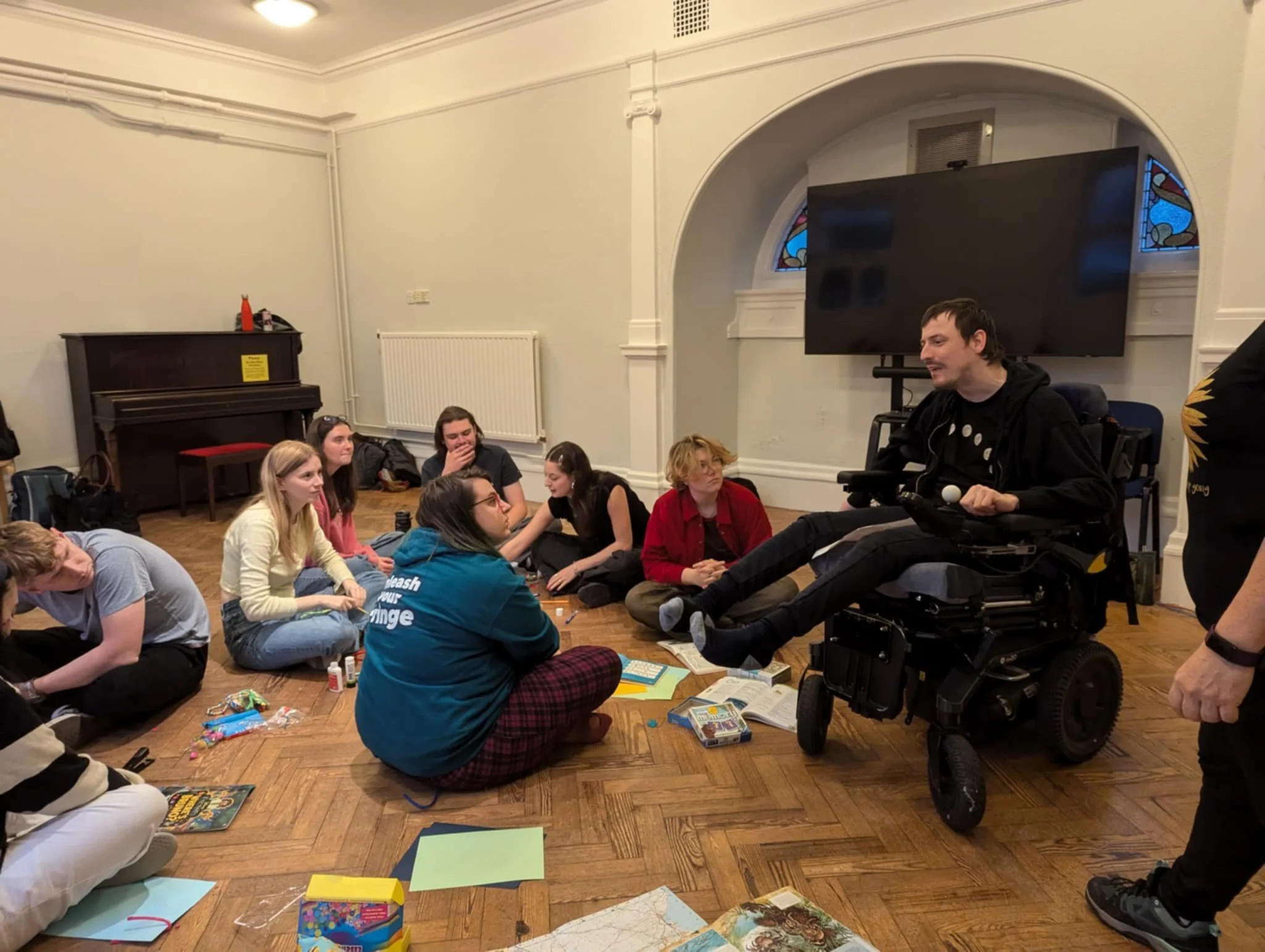 A group of student sitting on a floor around making equipment, they look up listening to a wheelchair user in black