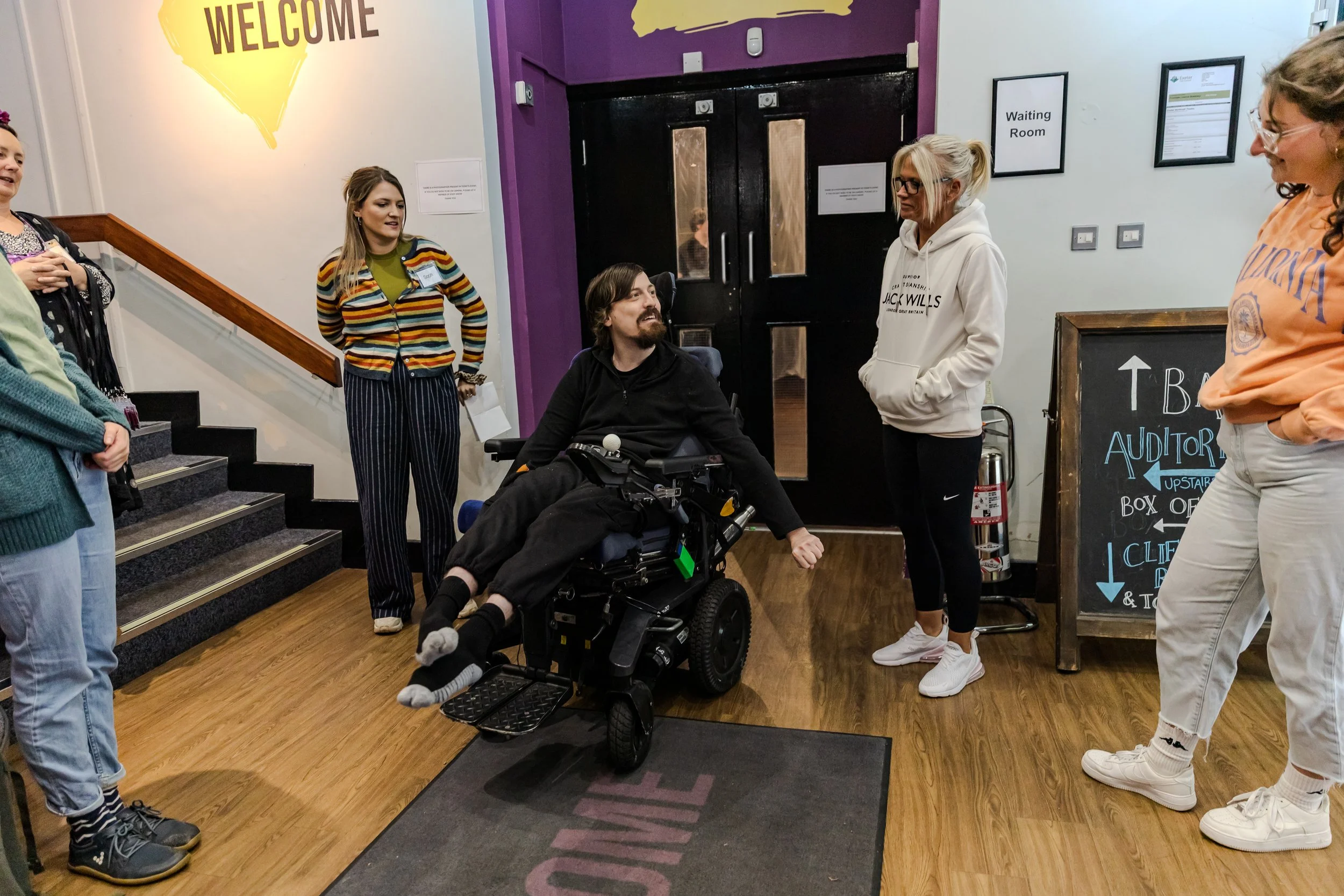 in a theatre foyer, a group of people listening in a circle to a performer wearing black with messy brown hair