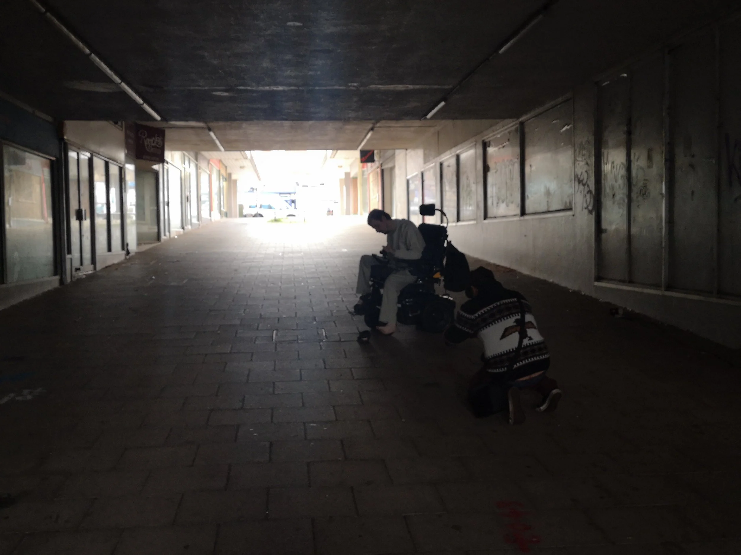 Hugh in white in his chair and Steve kneeling in a cardigan looking at  a sandal on the floor. They are surrounding by a concrete structure with graffiti and empty shop windows.