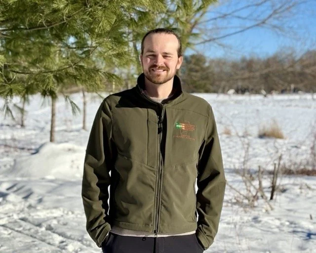 Adam Fisher is pictured standing in a sunny field under blue skies with snow in the background.