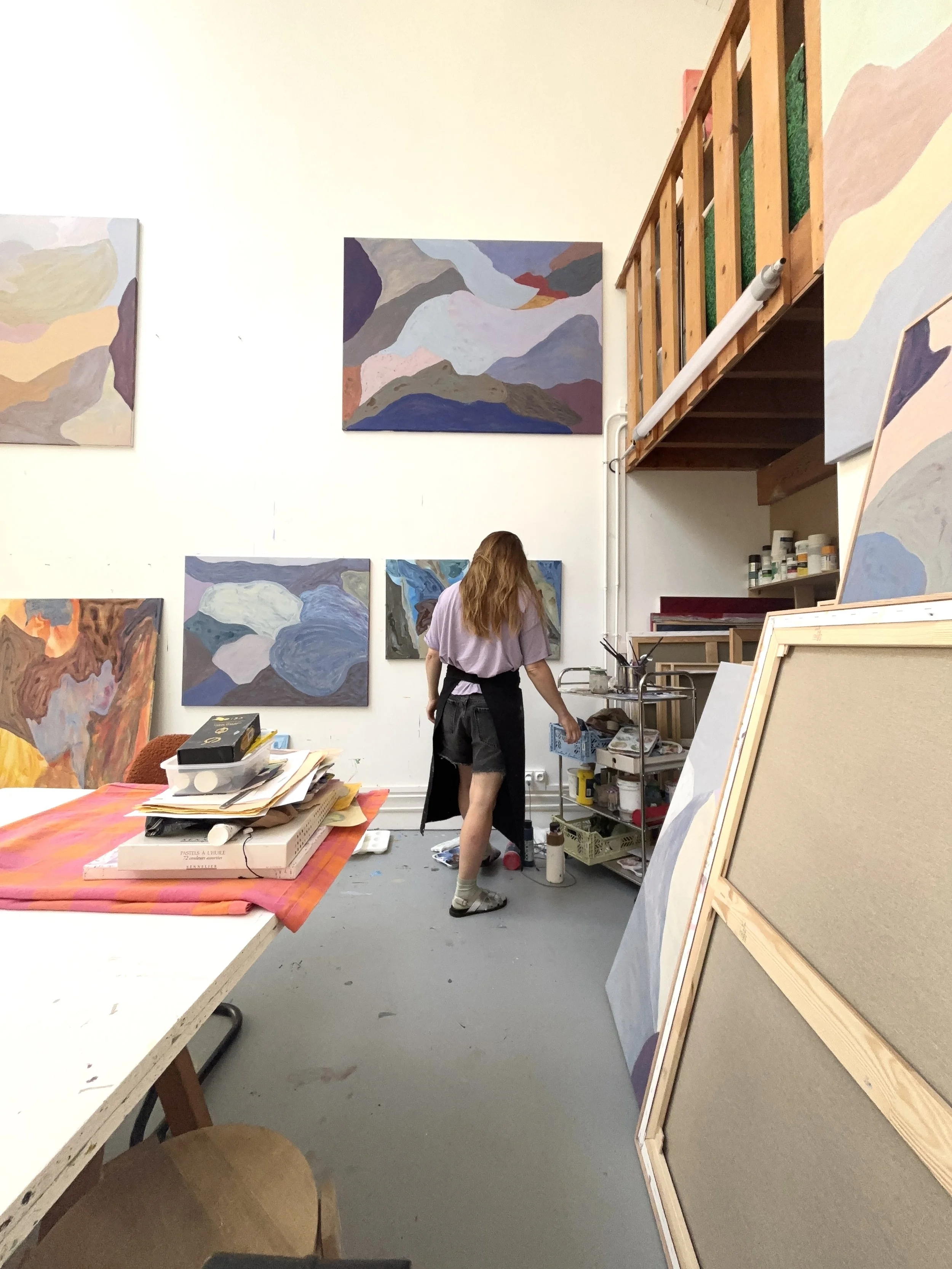 An artist's studio with colorful abstract mountain paintings on the walls, a woman with long, wavy hair, wearing a gray t-shirt, black shorts, and apron, working at a cart with art supplies.