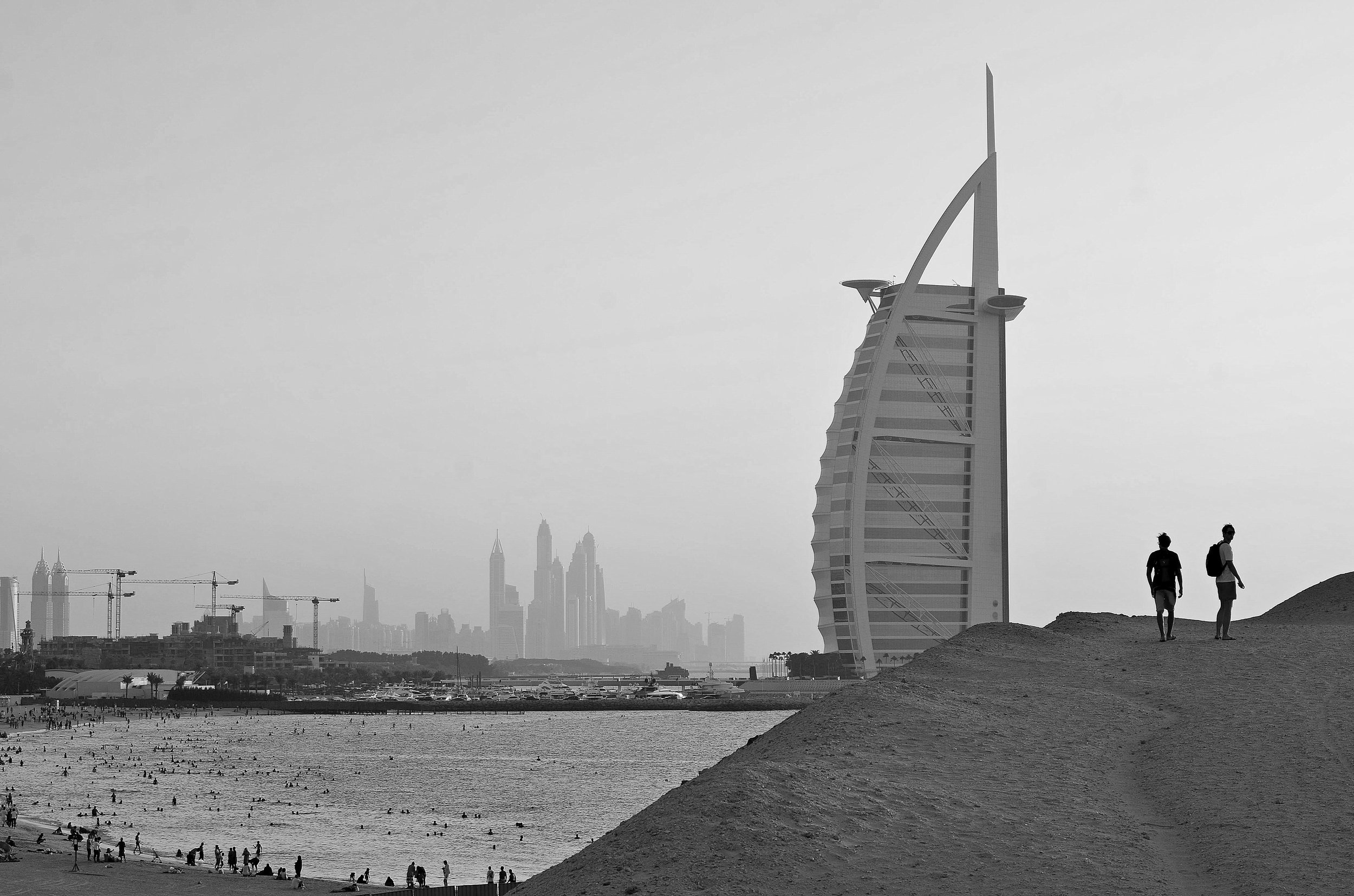 Black and white photo of the Dubai skyline featuring the Burj Al Arab hotel and a beach with many people, two people walking on a hill in the foreground.
