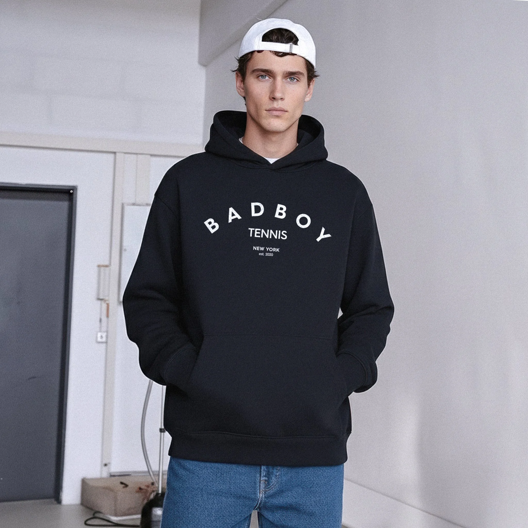 A young man wearing a black hoodie with the words "BADBOY TENNIS NEW YORK est. 2020" and a white baseball cap backwards. He has his hands in the hoodie pocket, standing indoors with a neutral expression.