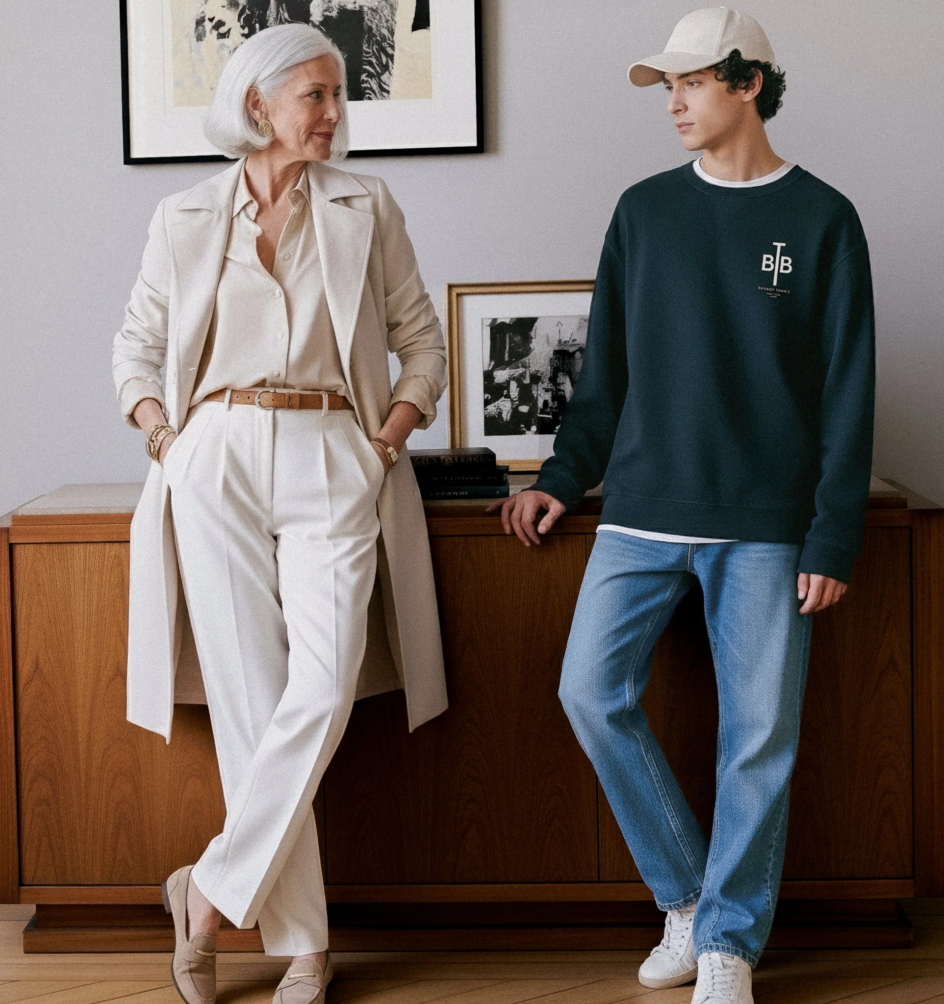 An older woman and a young man are standing in a room with modern artwork and photographs on the wall. The woman is dressed in a light-colored suit with a long coat, blouse, and high-waisted trousers, while the young man wears a dark sweatshirt, light blue jeans, a white cap, and white sneakers. They are engaged in conversation.