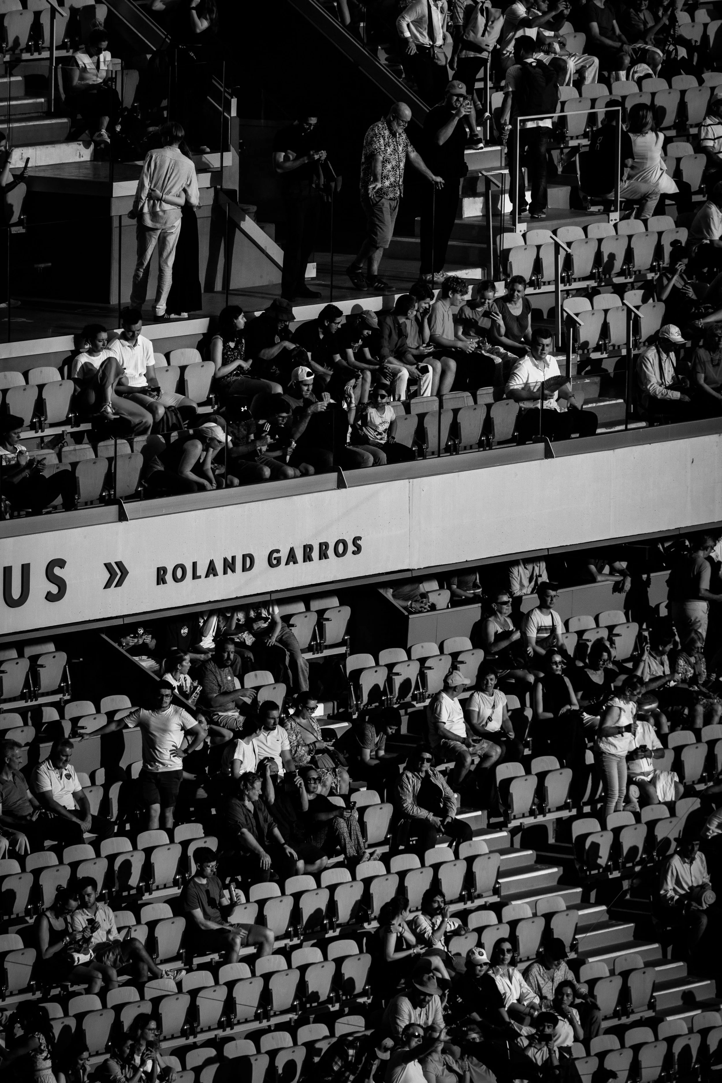 Crowd of people sitting and standing in stadium seats, some using phones, at Roland Garros tennis tournament, black and white photograph.