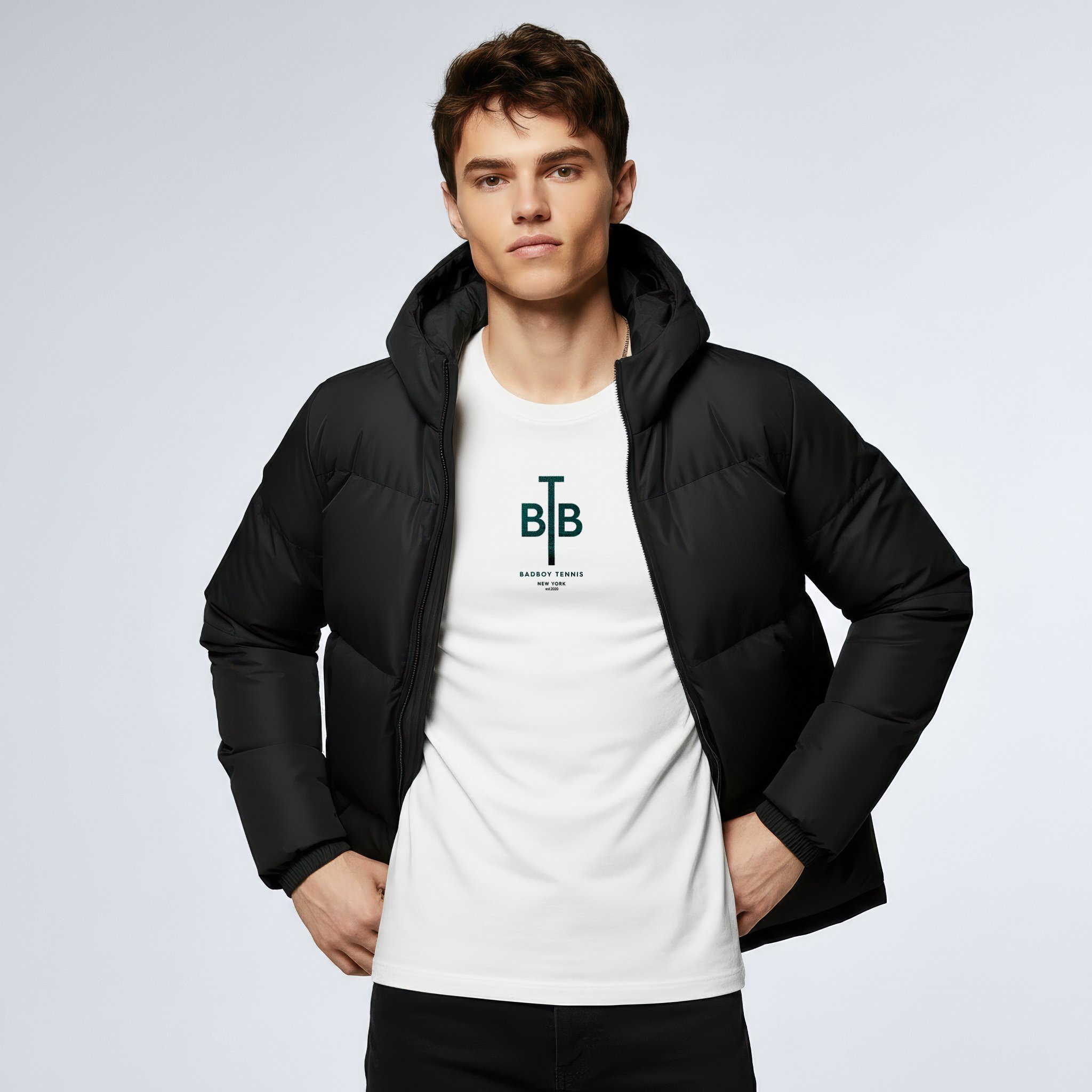 Young man wearing a black puffer jacket over a white T-shirt with a tennis logo, standing against a plain light gray background.