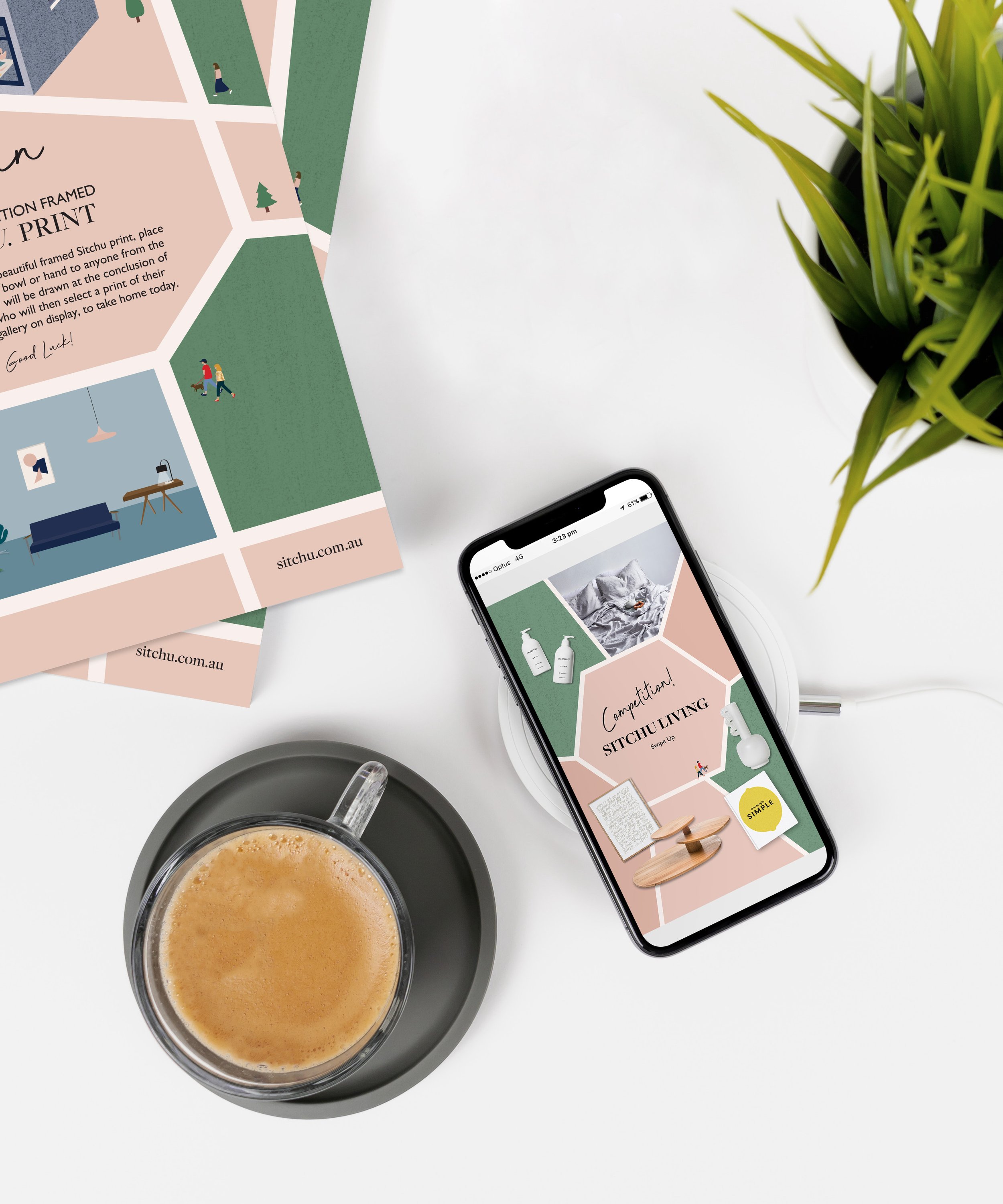 A white table with promotional plant-based living brochure, a smartphone displaying a design for a contest called 'Sitchu Living', a black saucer with a glass cup of coffee, a white wireless charging pad, and part of a green potted plant.