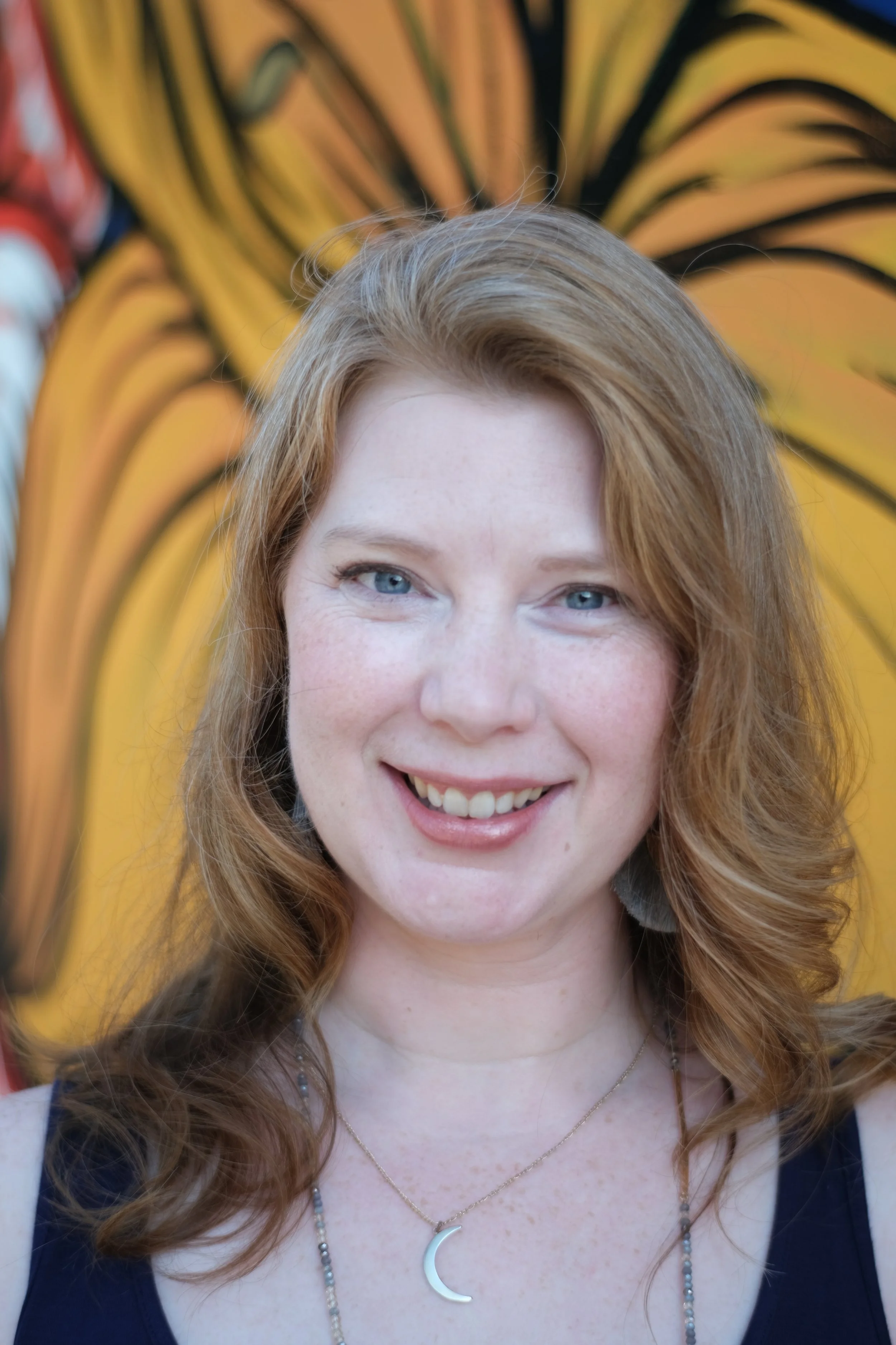 A young woman with red hair and blue eyes smiling in front of a yellow background with black swirling lines.
