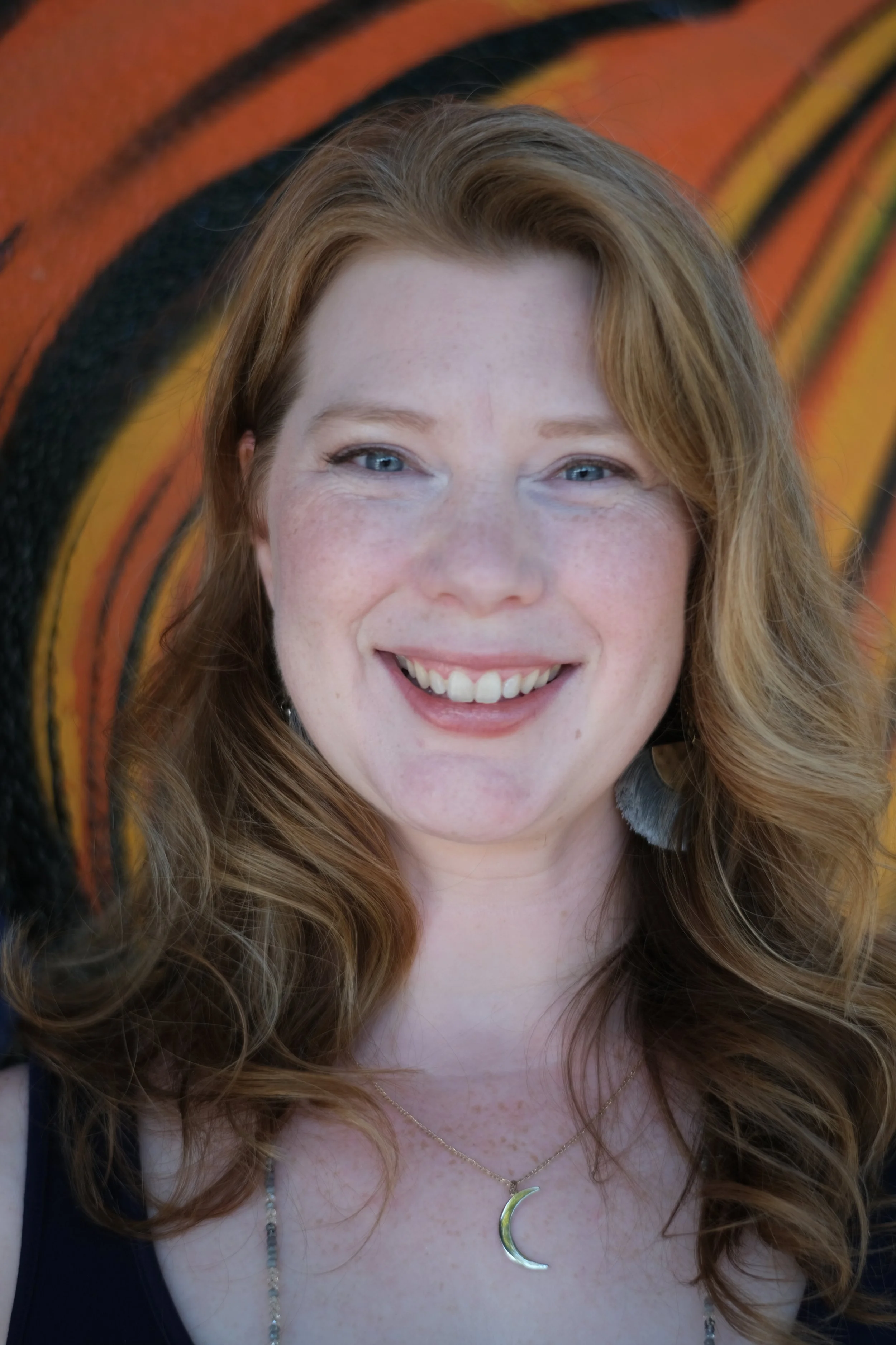 Close-up of a woman with long wavy red hair, blue eyes, and a big smile, wearing a gold crescent moon necklace and a beaded necklace. She is standing in front of an abstract, colorful background.