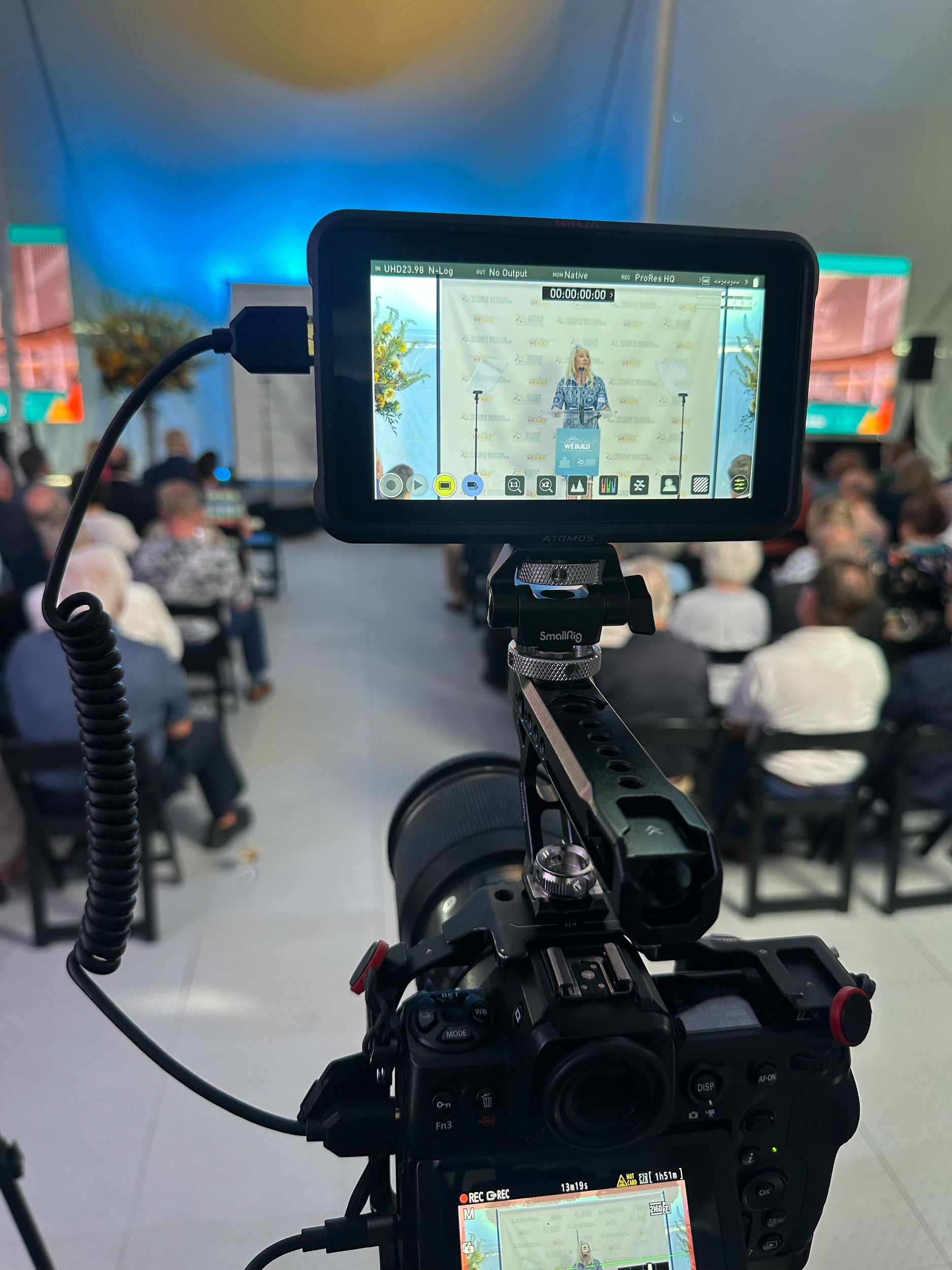 Camera filming a woman giving a speech at a podium during a conference, with an audience seated in front of her.