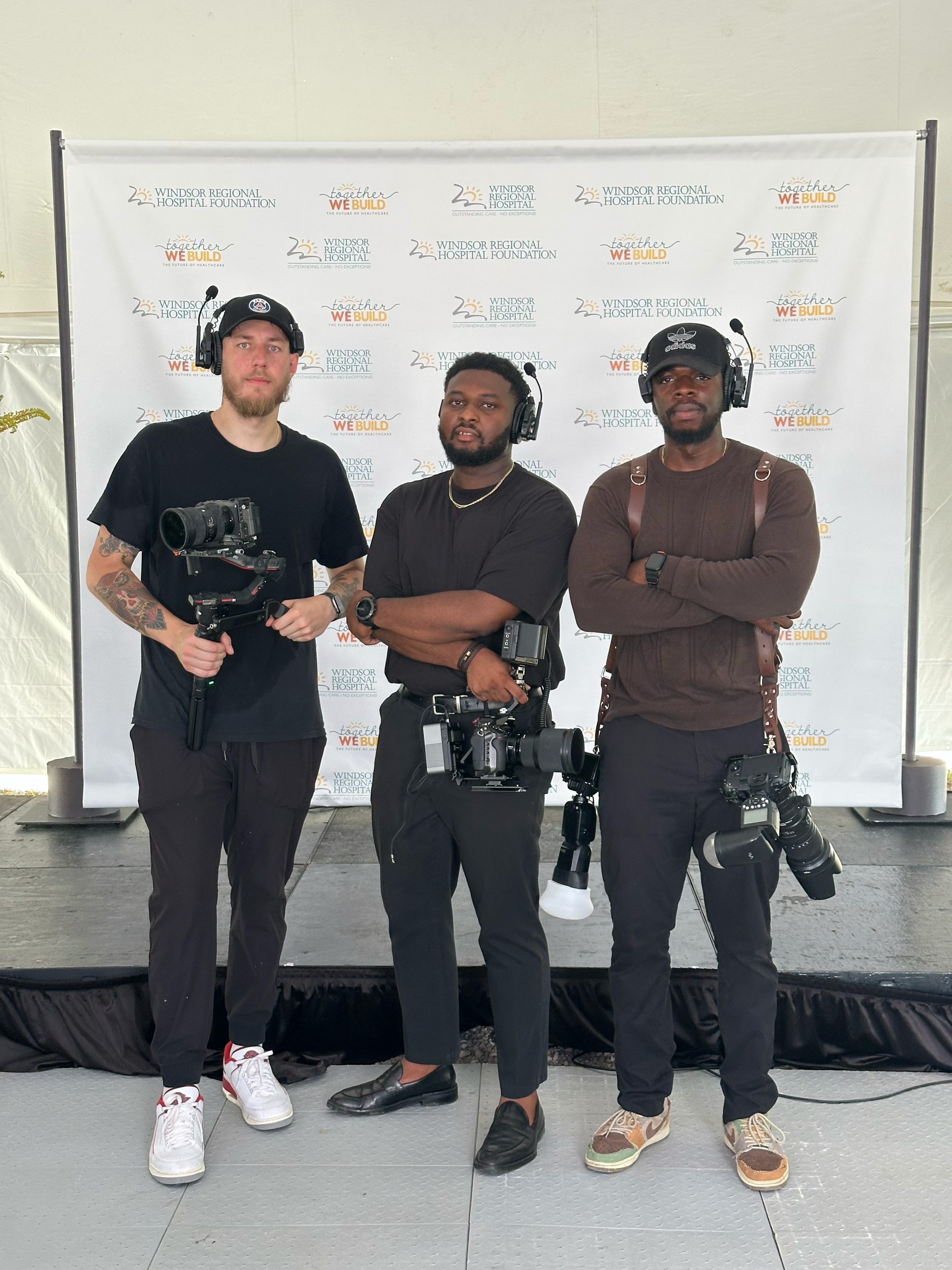 Three men standing in front of a backdrop with logos, holding cameras and wearing headsets.