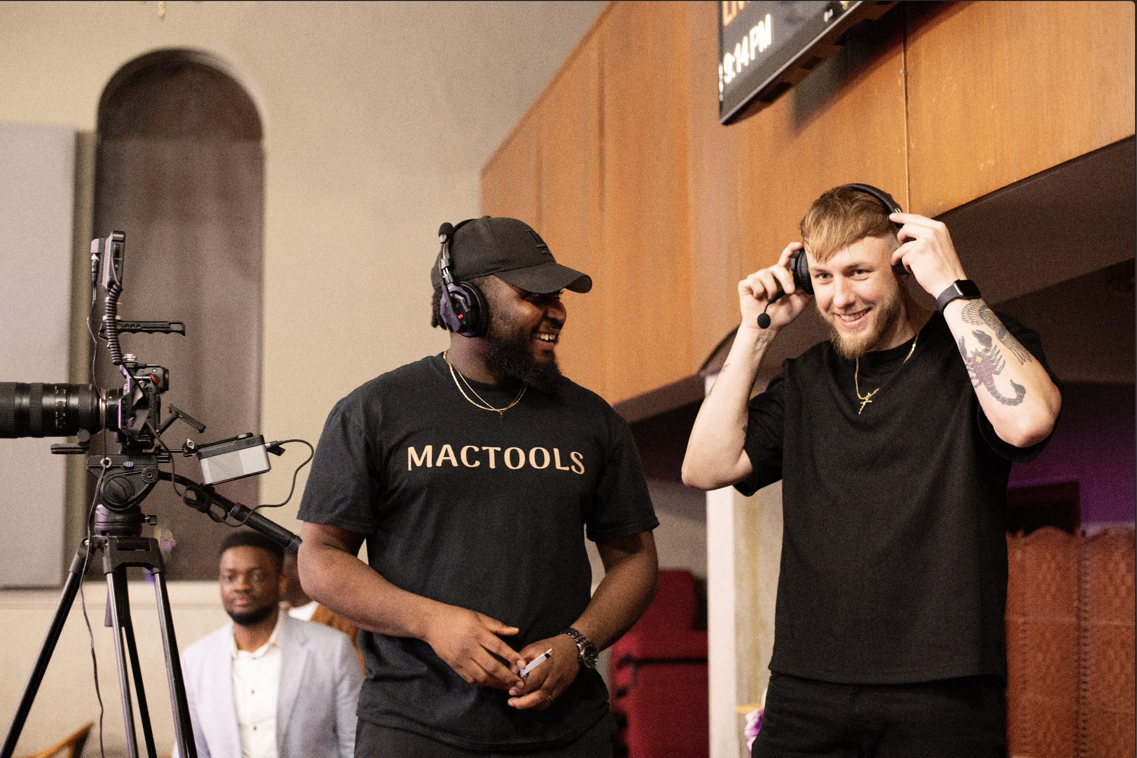 Two men smiling and wearing headphones at an indoor event, one man is wearing a black 'MACTOOLS' t-shirt, and the other has tattoos on his arms and is adjusting his headphones.
