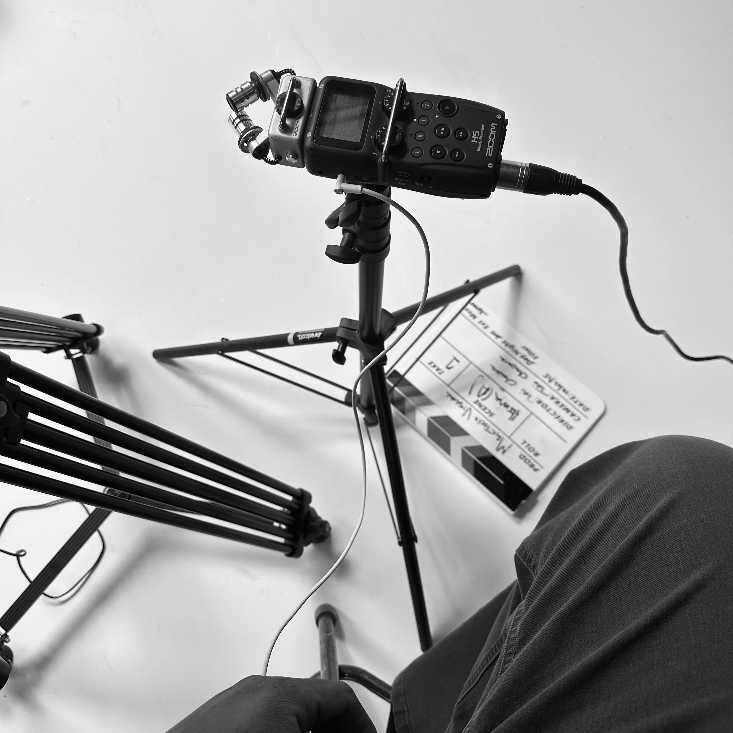 Black and white photo showing a camera mounted on a tripod, video lighting equipment, and a clapperboard on the floor, with part of a person's leg and hand visible.