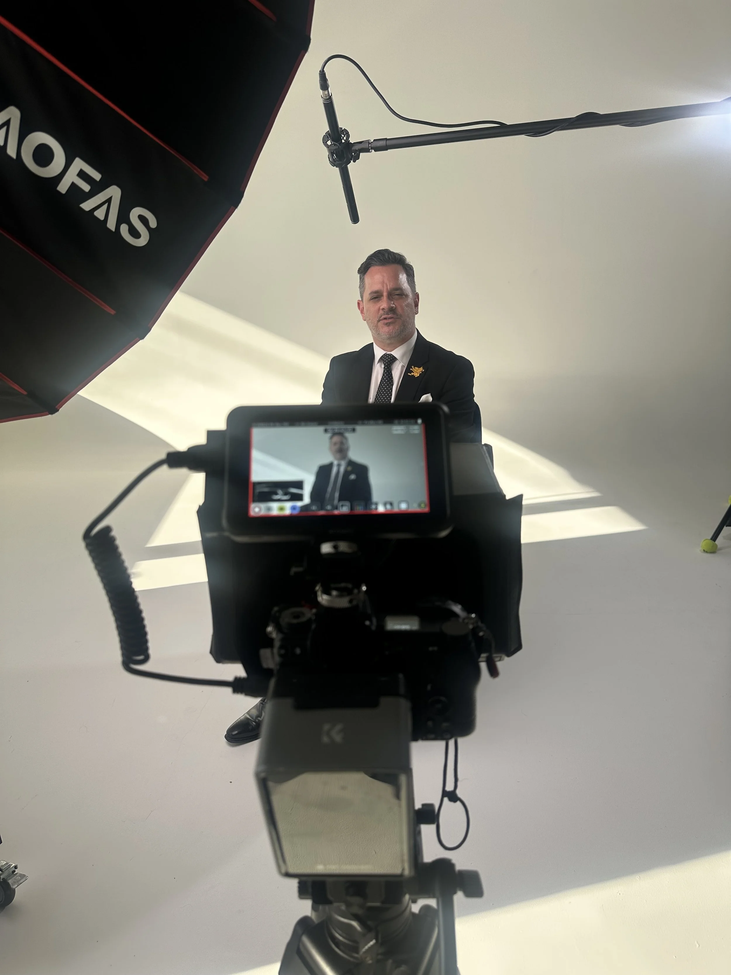 A man in a suit and tie is being filmed in front of a plain light-colored background, with studio equipment including a camera and lighting setup visible.