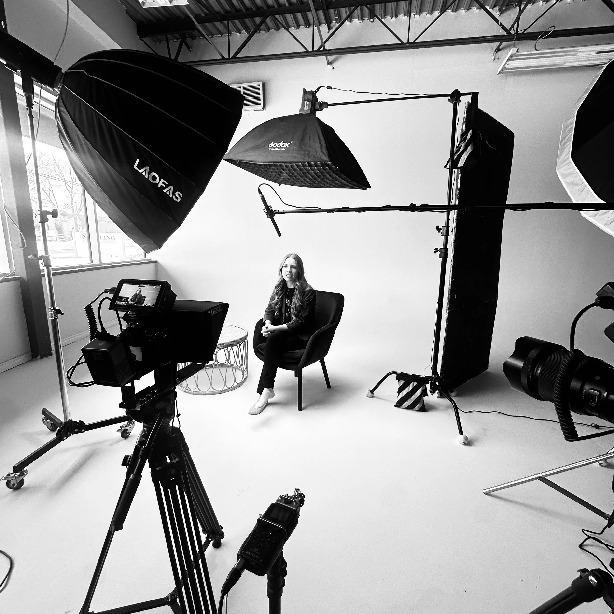 Black and white photo of a woman sitting on a chair in a photo studio with lighting and camera equipment around her, a white backdrop behind her, and a window on the left side.