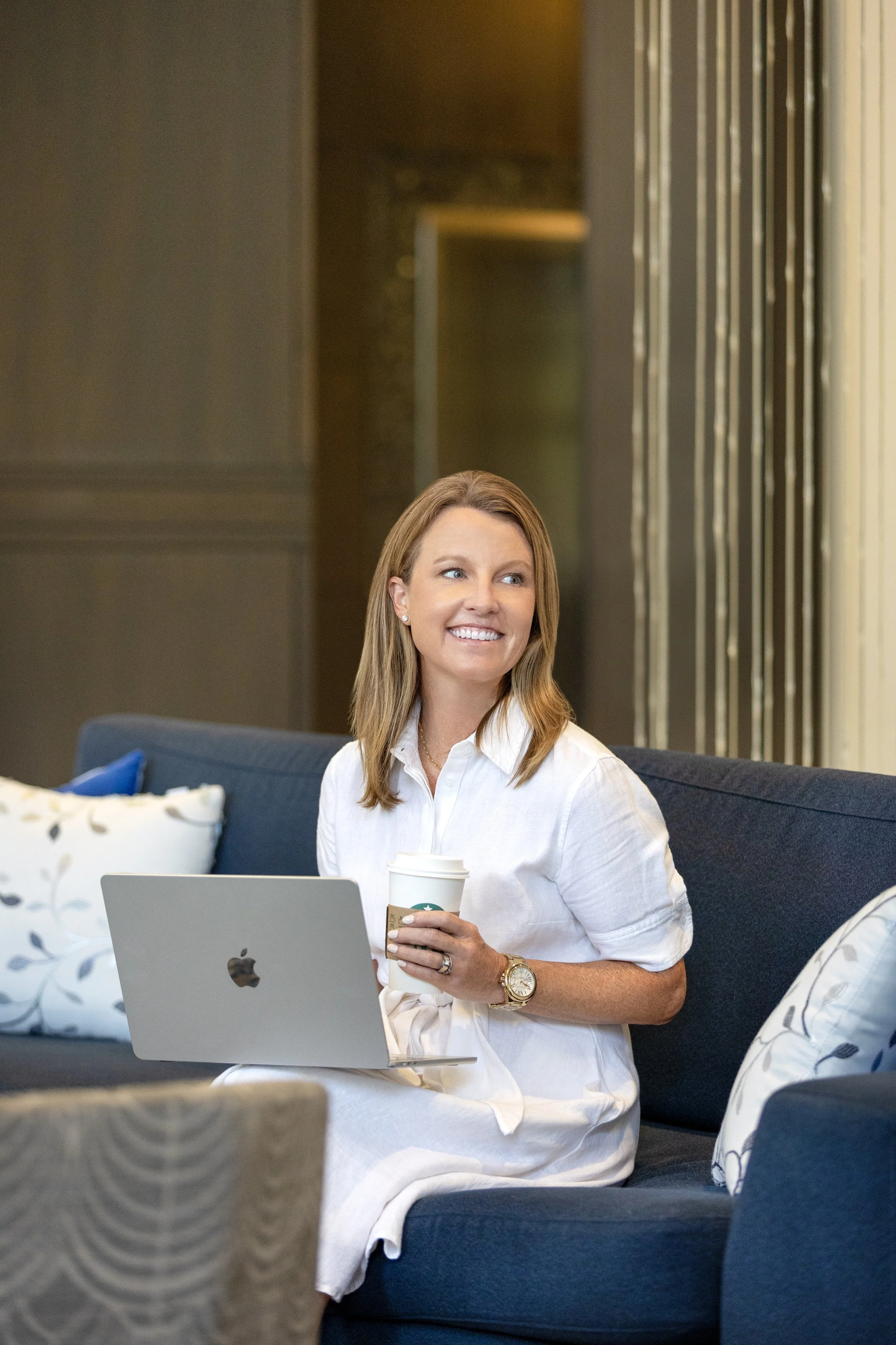 A woman with light brown hair, wearing a white dress and a gold watch, is sitting on a blue sofa, holding a Starbucks coffee cup, with a silver laptop in front of her. She is smiling and looking to her right, in a modern indoor setting with decorative pillows and a background with vertical metallic panels.