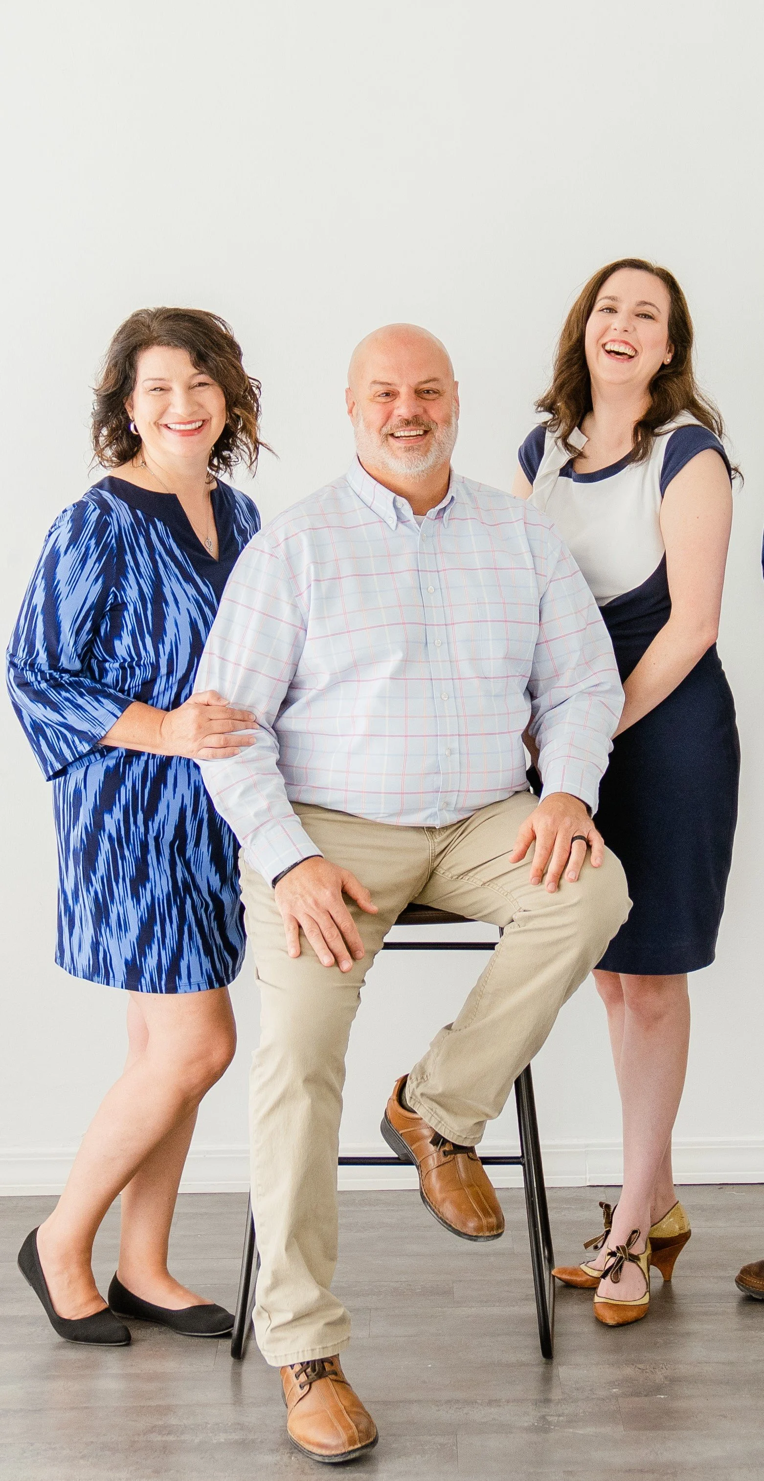 Three people posing indoors; one seated on a stool with two standing beside, all smiling and dressed in casual attire.