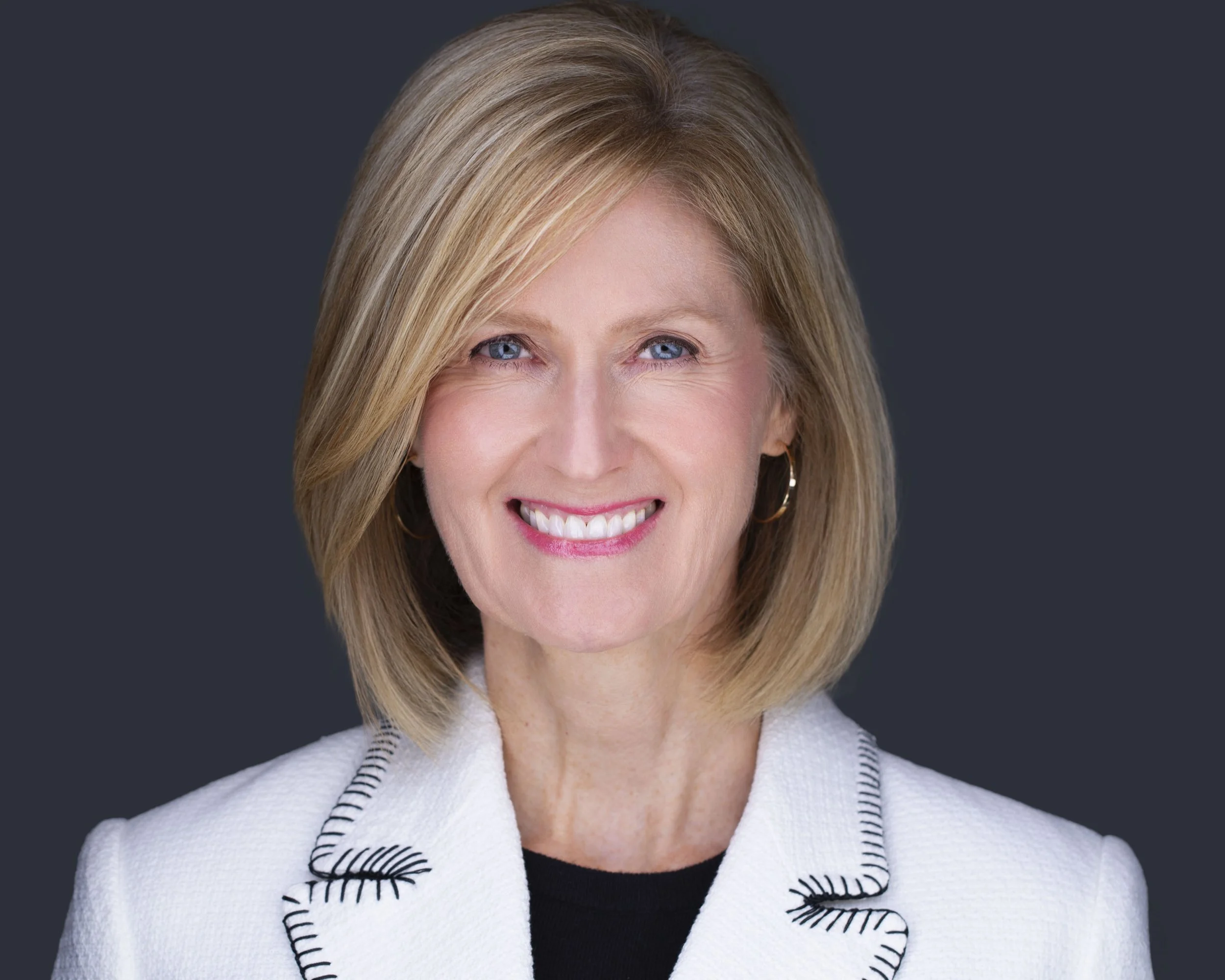 Professional headshots Dallas TX – executive portrait of woman in white jacket with shoulder-length blonde hair, smiling confidently in Dallas headshot studio with dark background.