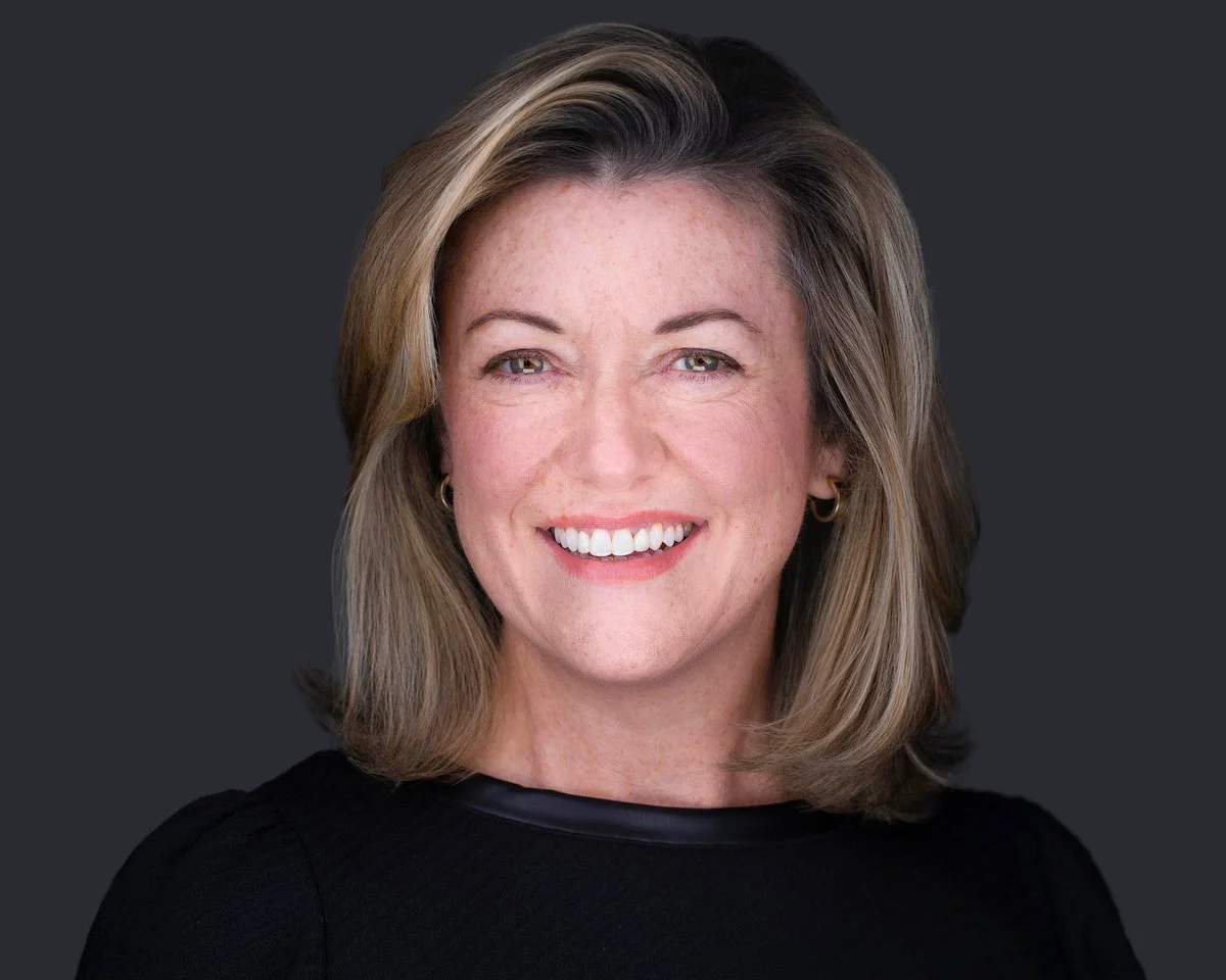 Professional headshot of Katie smiling, wearing black against a charcoal background.