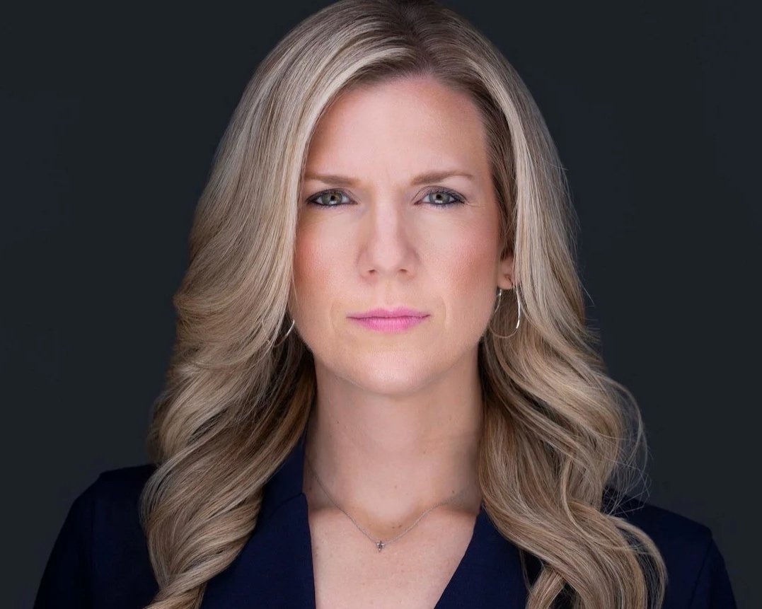 Woman with long blonde hair wearing a dark blazer and necklace against a dark background.