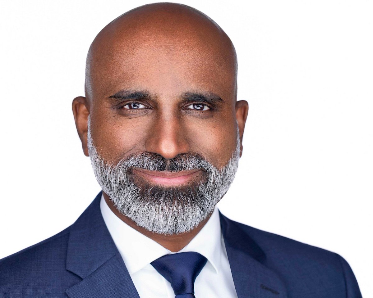 Man with a gray beard wearing a navy suit and tie against a white background.