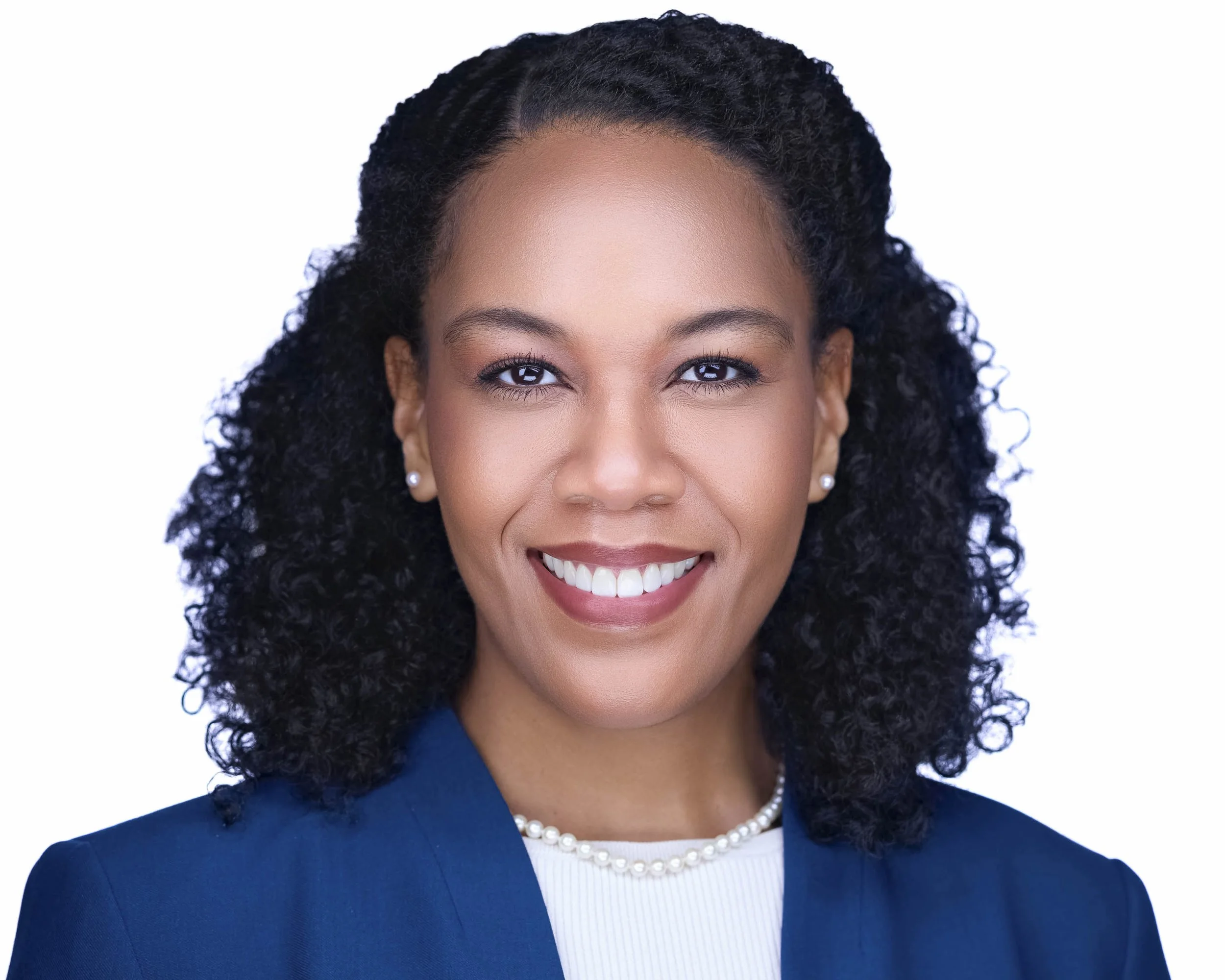 Professional headshots Dallas TX – business portrait of woman with naturally curly hair pulled back, wearing a navy blazer over a white blouse with a pearl necklace, smiling in Dallas by Godoy Shots headshot studio with bright background.
