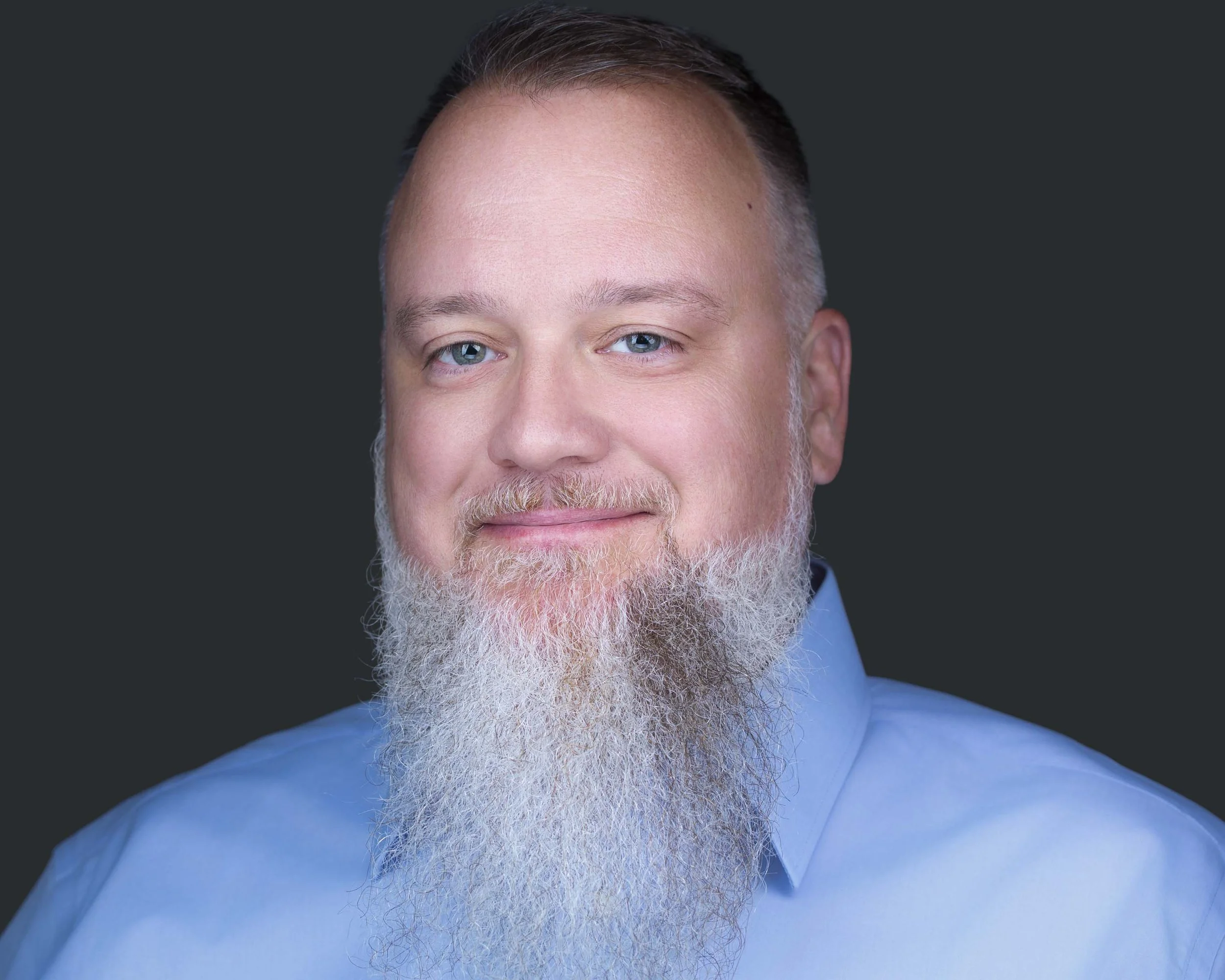 Portrait of a man with full white beard, wearing a light blue dress shirt, smiling with a relaxed expression in Dallas by Godoy Shots headshot studio with dark background