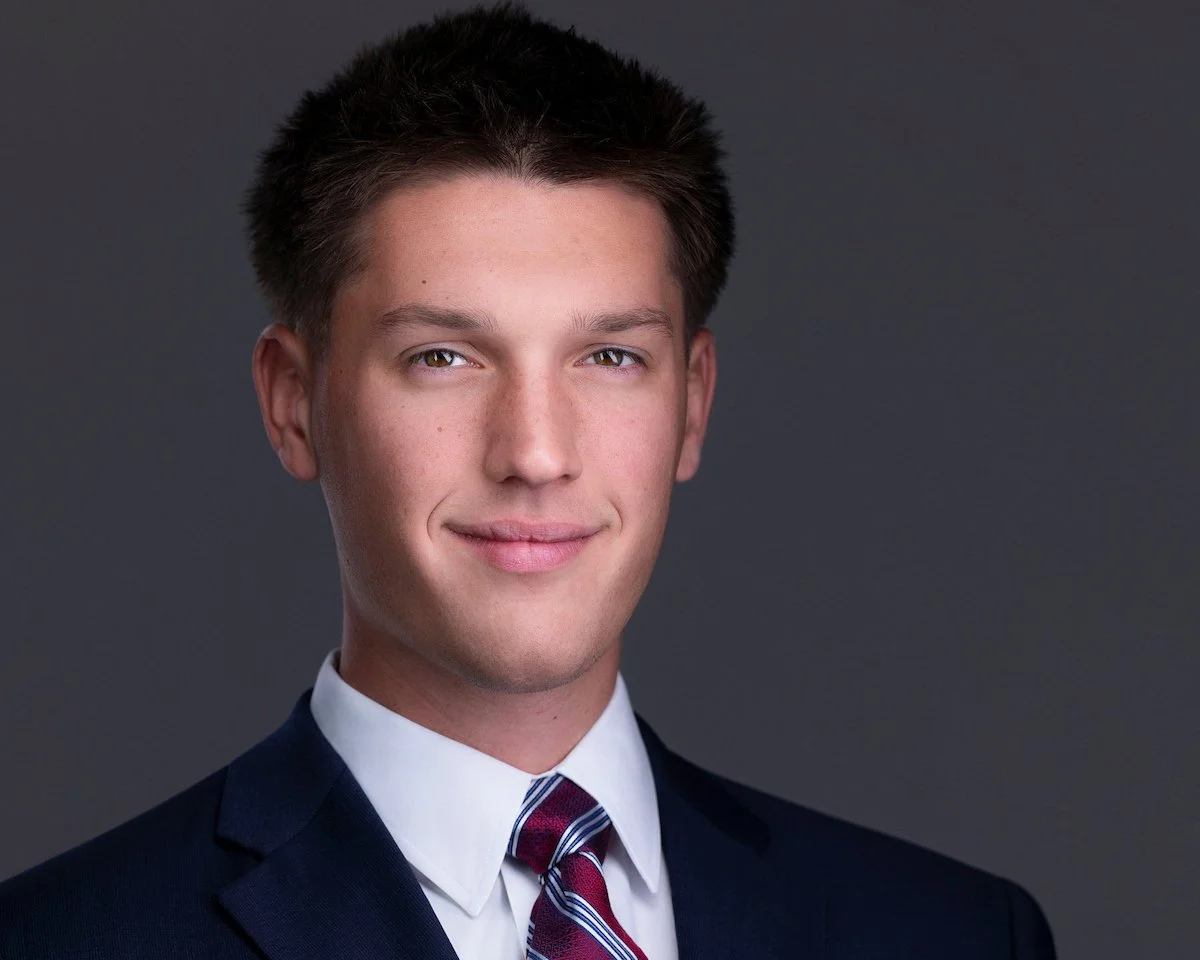 Man in a dark suit and striped tie against a charcoal background.