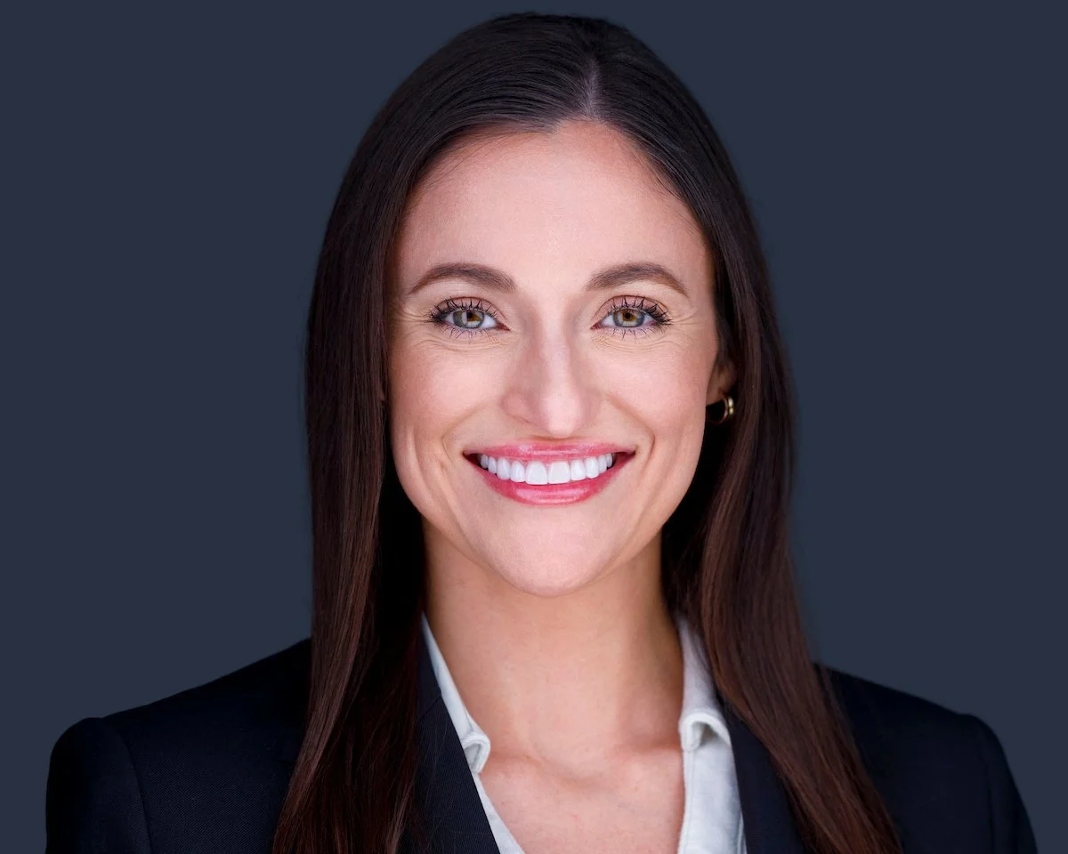 Smiling woman in a dark blazer and white collared shirt against a dark background.