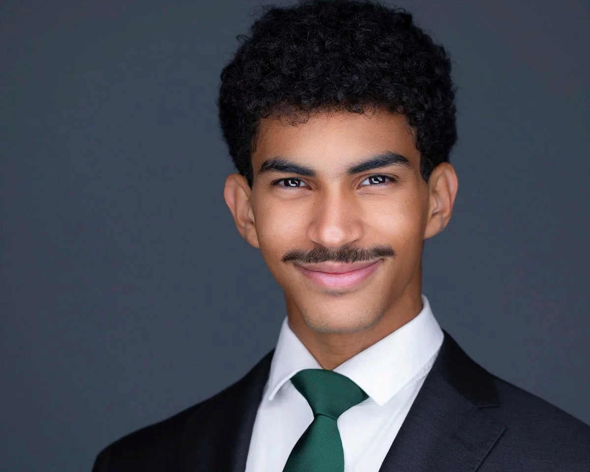 Smiling man with curly hair and a mustache wearing a dark suit, white shirt, and green tie against a gray background.