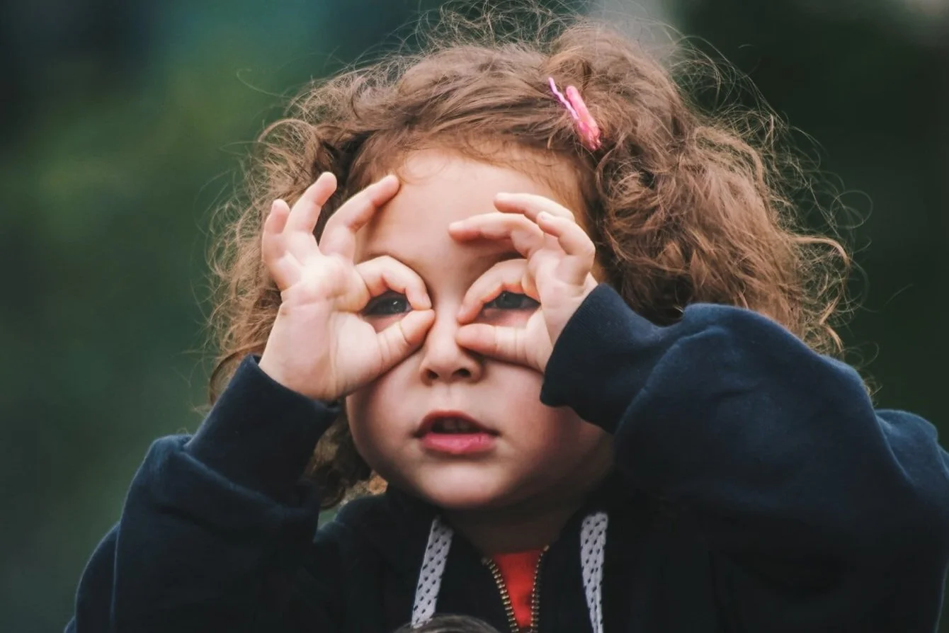 A young girl with curly hair and a pink hair clip making binoculars with her hands around her eyes outdoors.